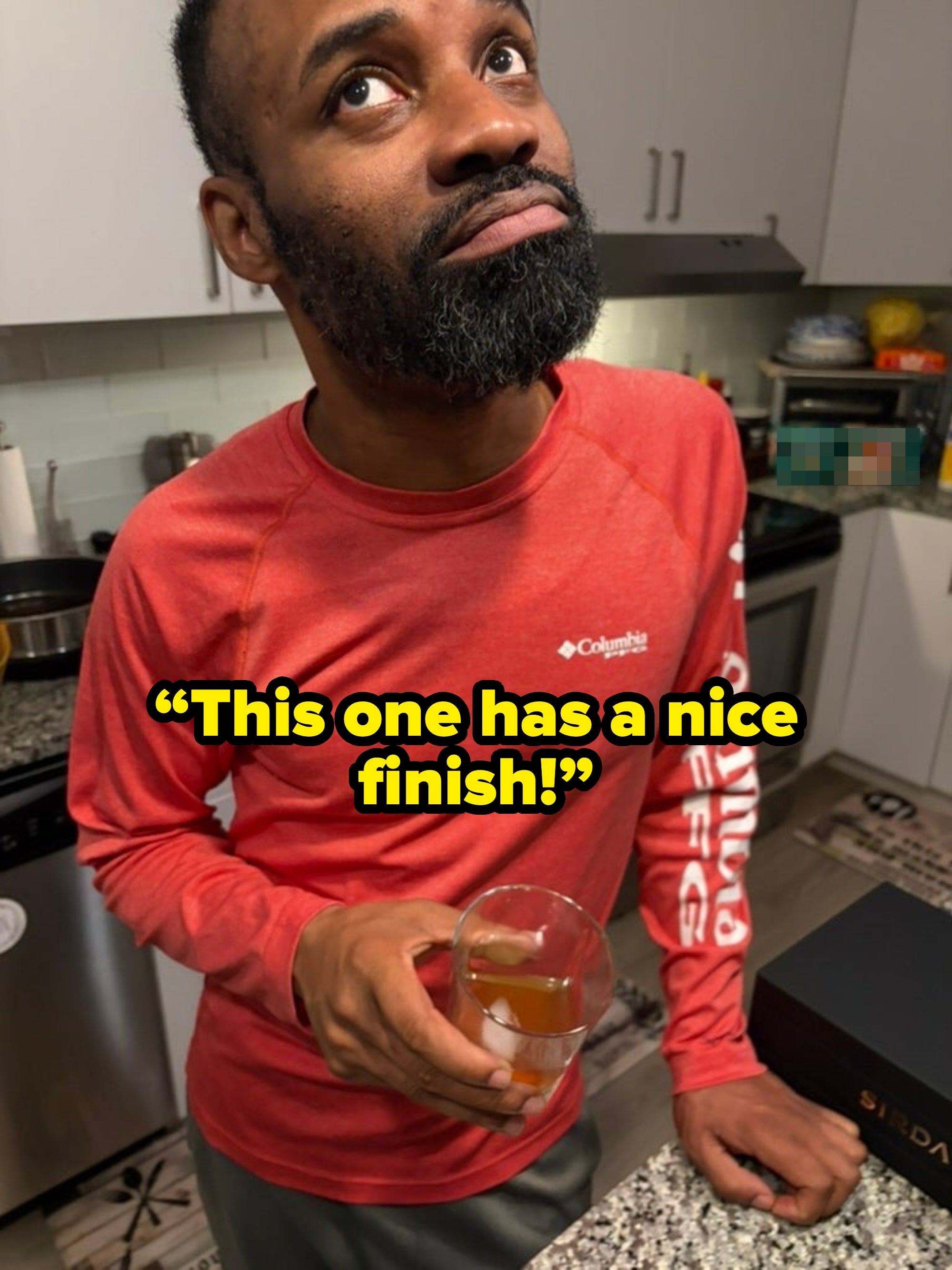 Person in a kitchen holds a glass of brown beverage, wearing a casual long-sleeve shirt, looking upwards thoughtfully