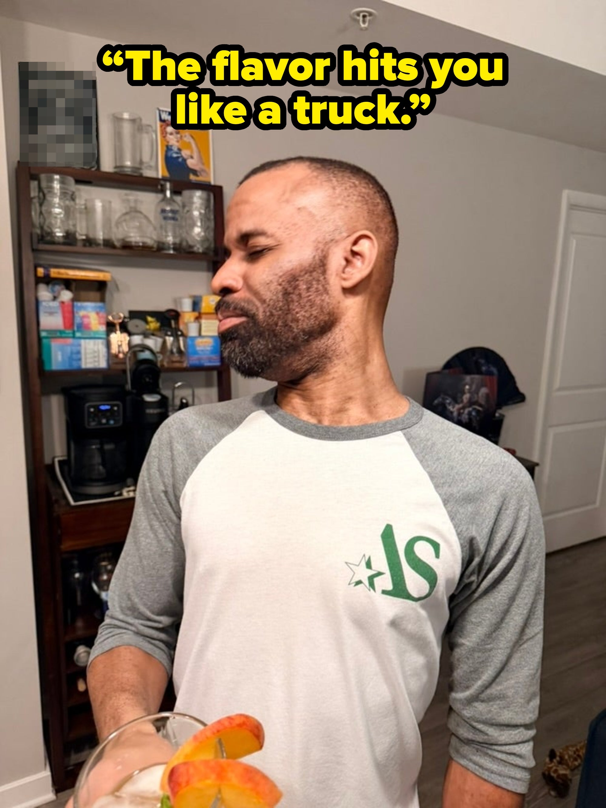 A man in a casual raglan shirt holds a cocktail with fruit garnish, standing in a kitchen with shelves and a coffee maker in the background