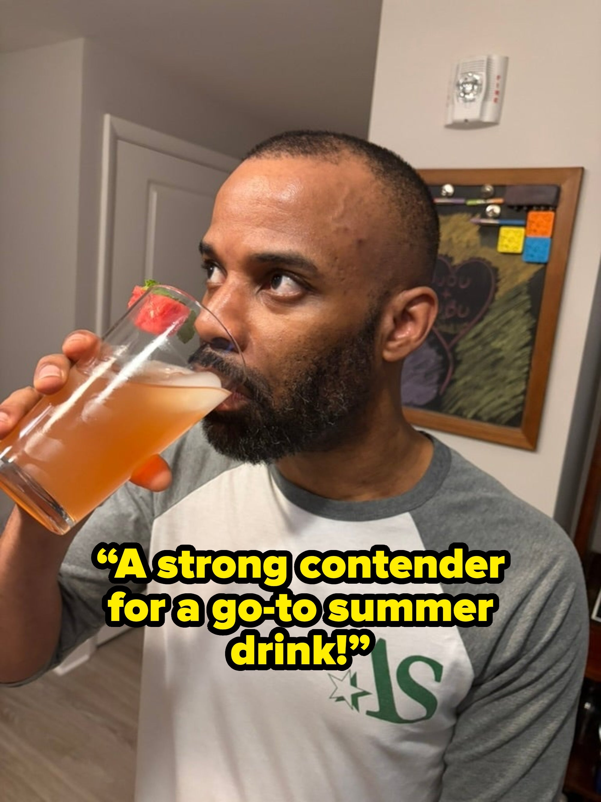 Person drinking a beverage from a glass with a strawberry garnish, standing in a kitchen