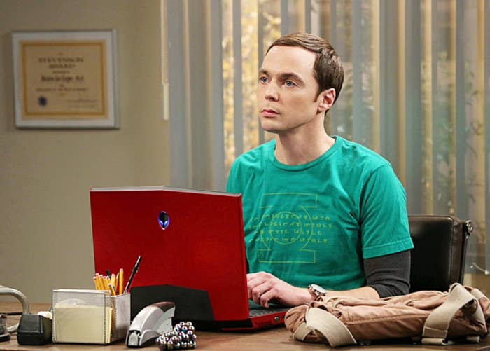 Person with short hair wearing a T-shirt, sitting at a desk with a red laptop, surrounded by office items