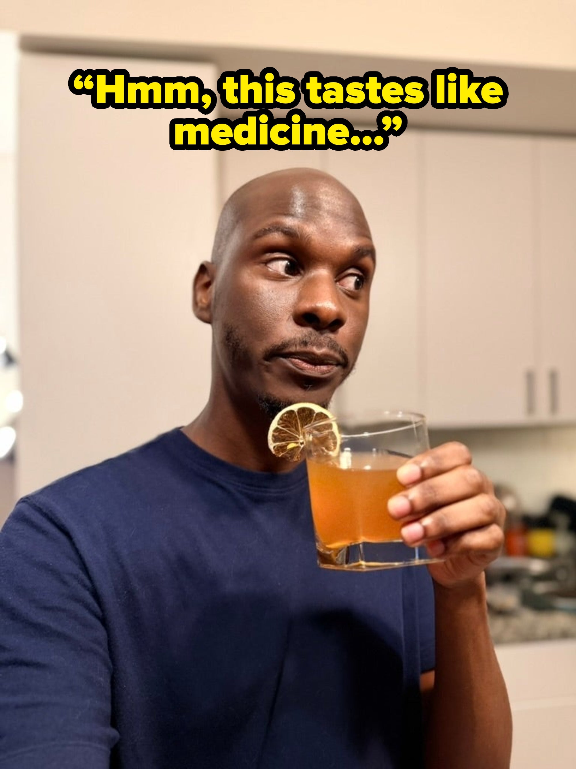 Man drinking a citrus-garnished beverage in a kitchen, looking contemplative