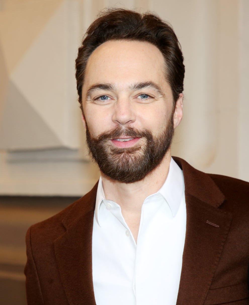 A man with a beard is wearing a white shirt and brown blazer, smiling at the camera