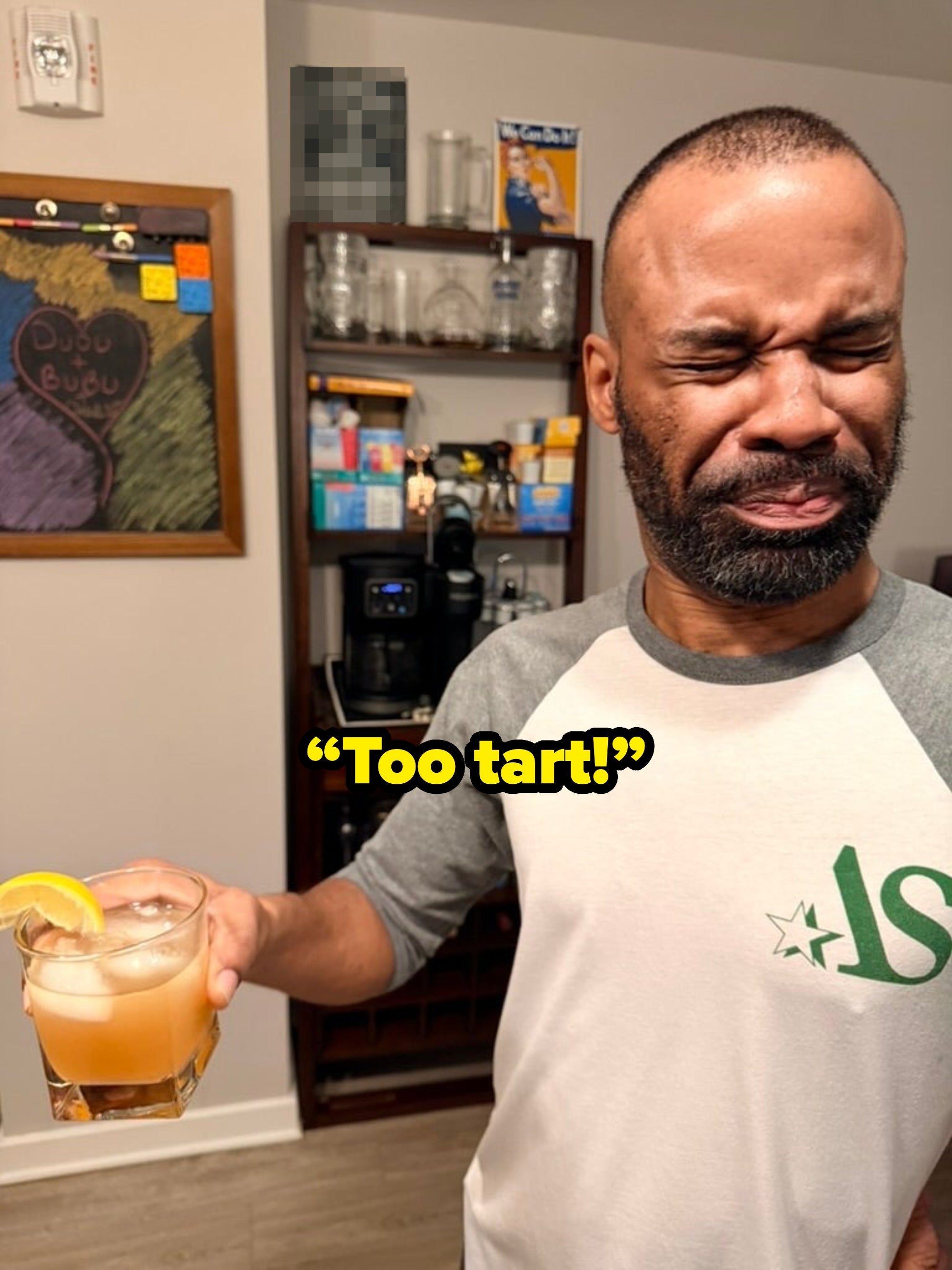 Person grimaces while holding a cocktail with an orange slice, suggesting a strong or sour taste. Behind them are kitchen shelves and a chalkboard