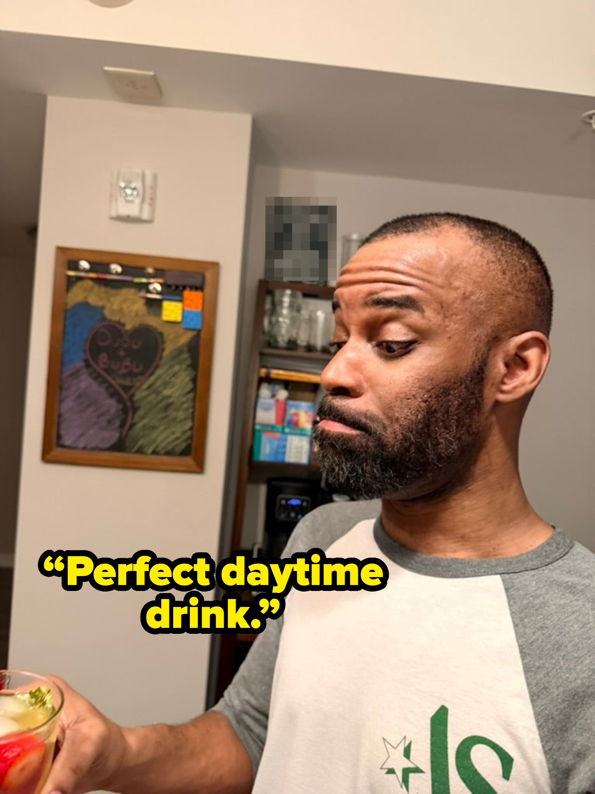 A person in a casual shirt looks curiously at a beverage in their hand, standing in a kitchen with a chalkboard in the background