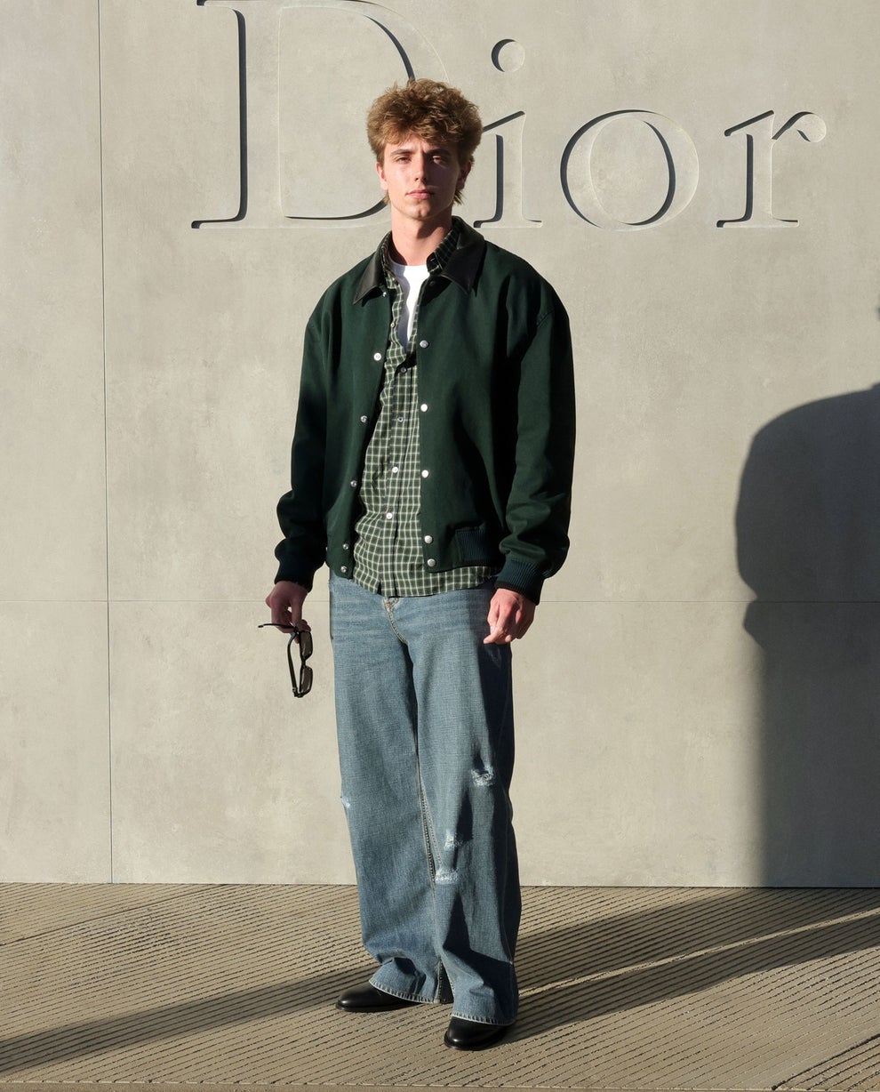 Person wearing a casual green jacket over a checkered shirt and loose jeans, holding sunglasses, stands in front of a wall with "Dior" in the background
