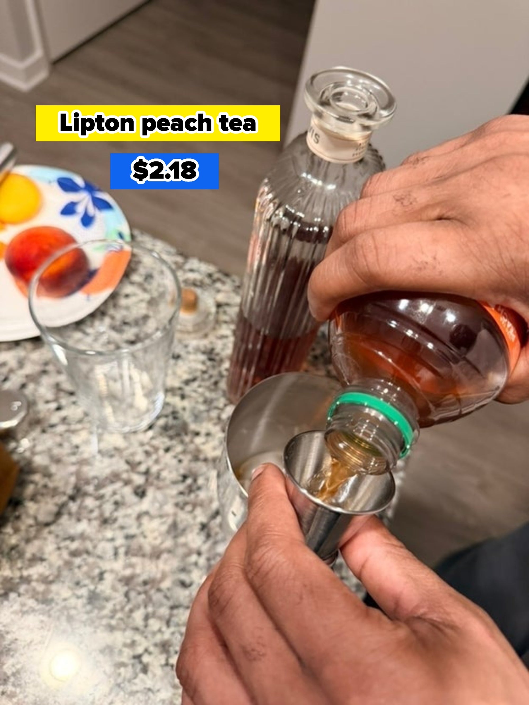 A person pours liquor from a bottle into a jigger over a countertop. A glass and a plate with sliced fruit are nearby