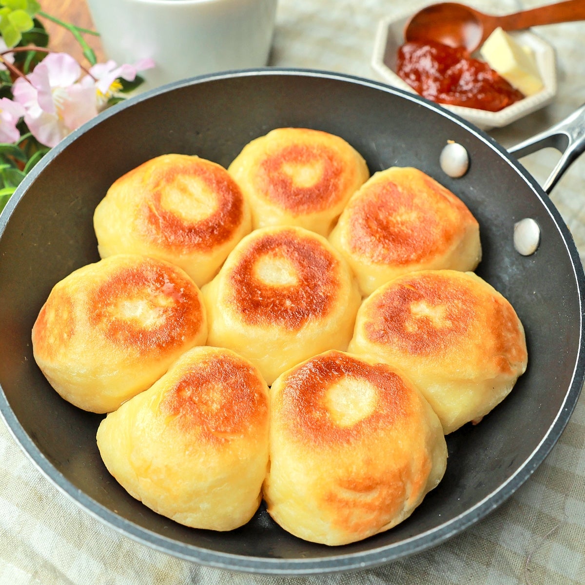 フライパンで焼かれた金色の焼き立てパンが8個あります。背景にはジャムとバターが並んでいます。