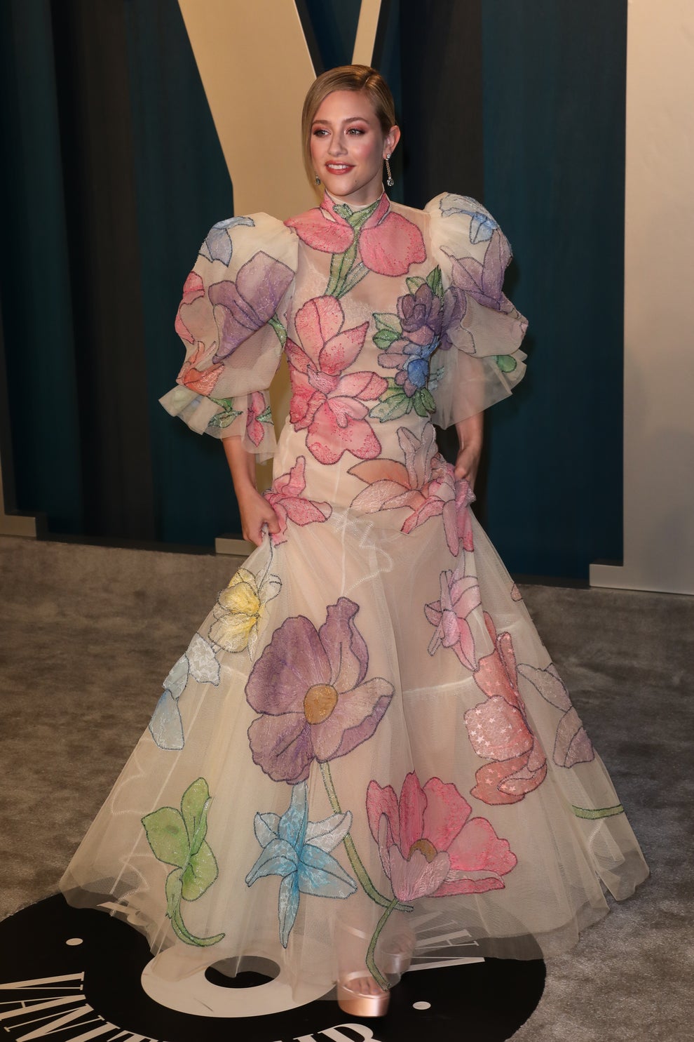 Lili Reinhart in voluminous, floral-patterned gown with sheer fabric, standing on a carpet at an event