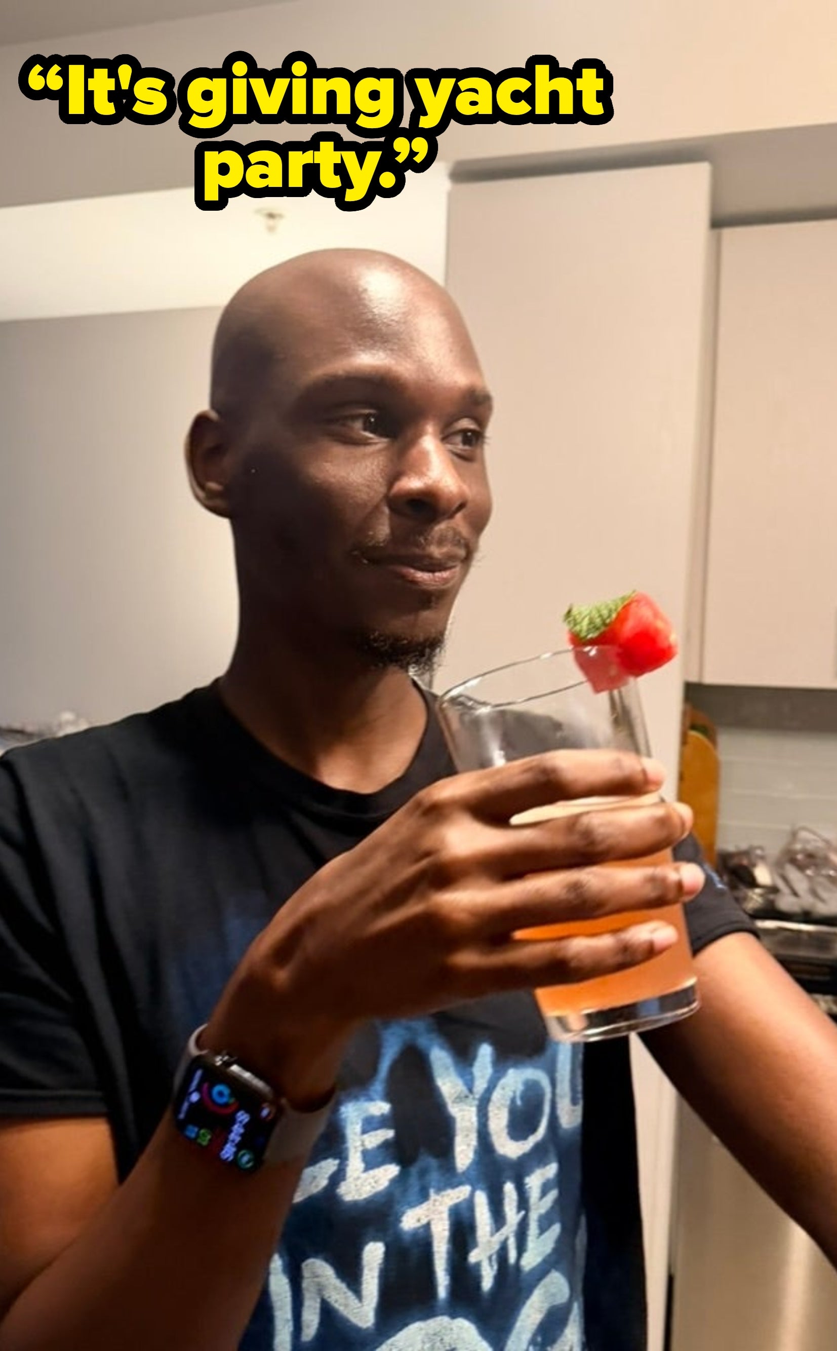 Person holding a cocktail garnished with watermelon and mint in a kitchen, wearing a casual t-shirt and smartwatch