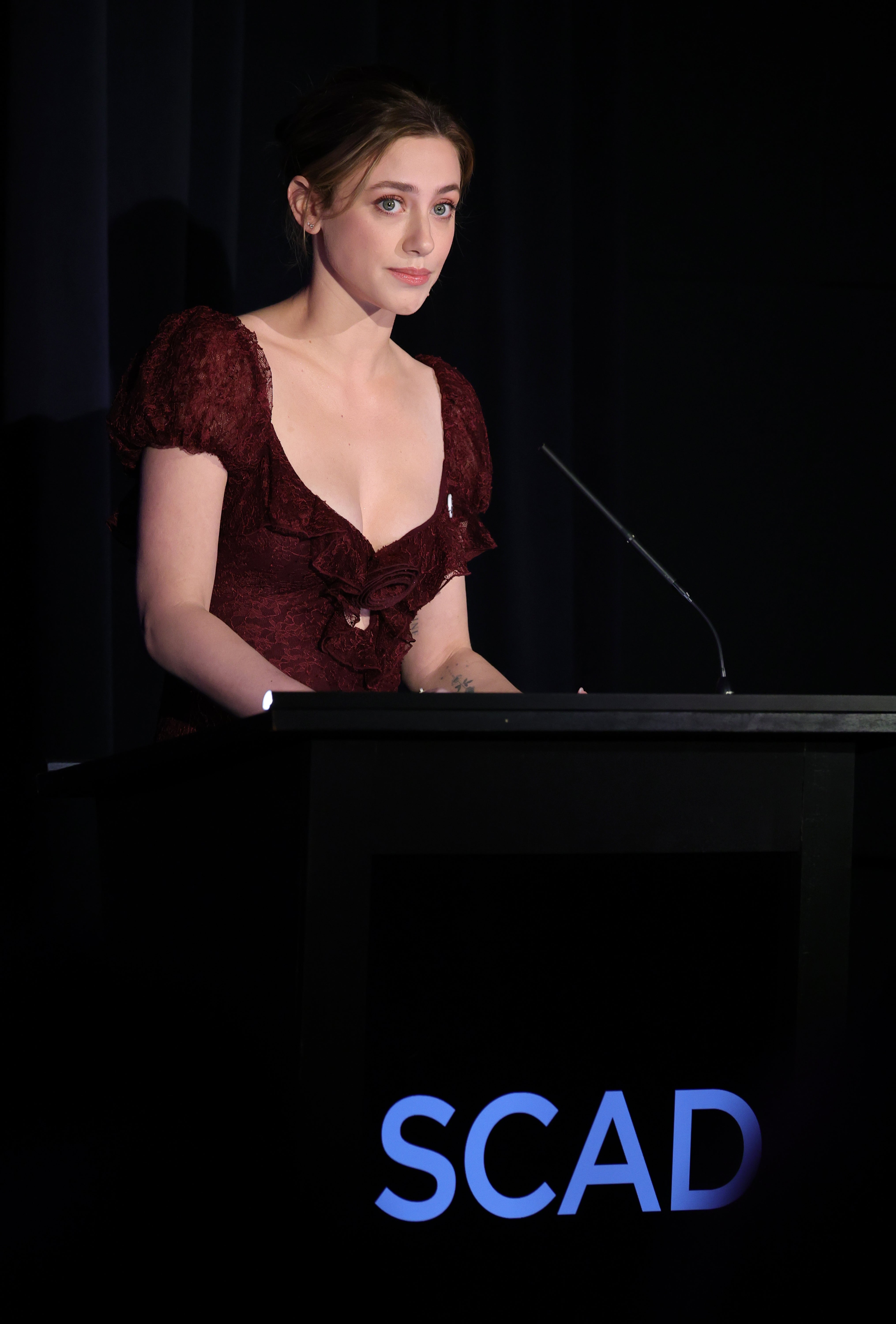 Lili Reinhart at podium wearing an elegant dress with ruffled details, speaking at a SCAD event