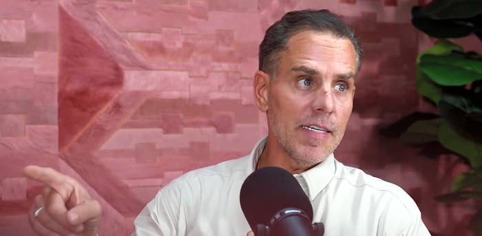 Person speaking into a microphone, gesturing to the side, seated in front of a wall with a geometric pattern