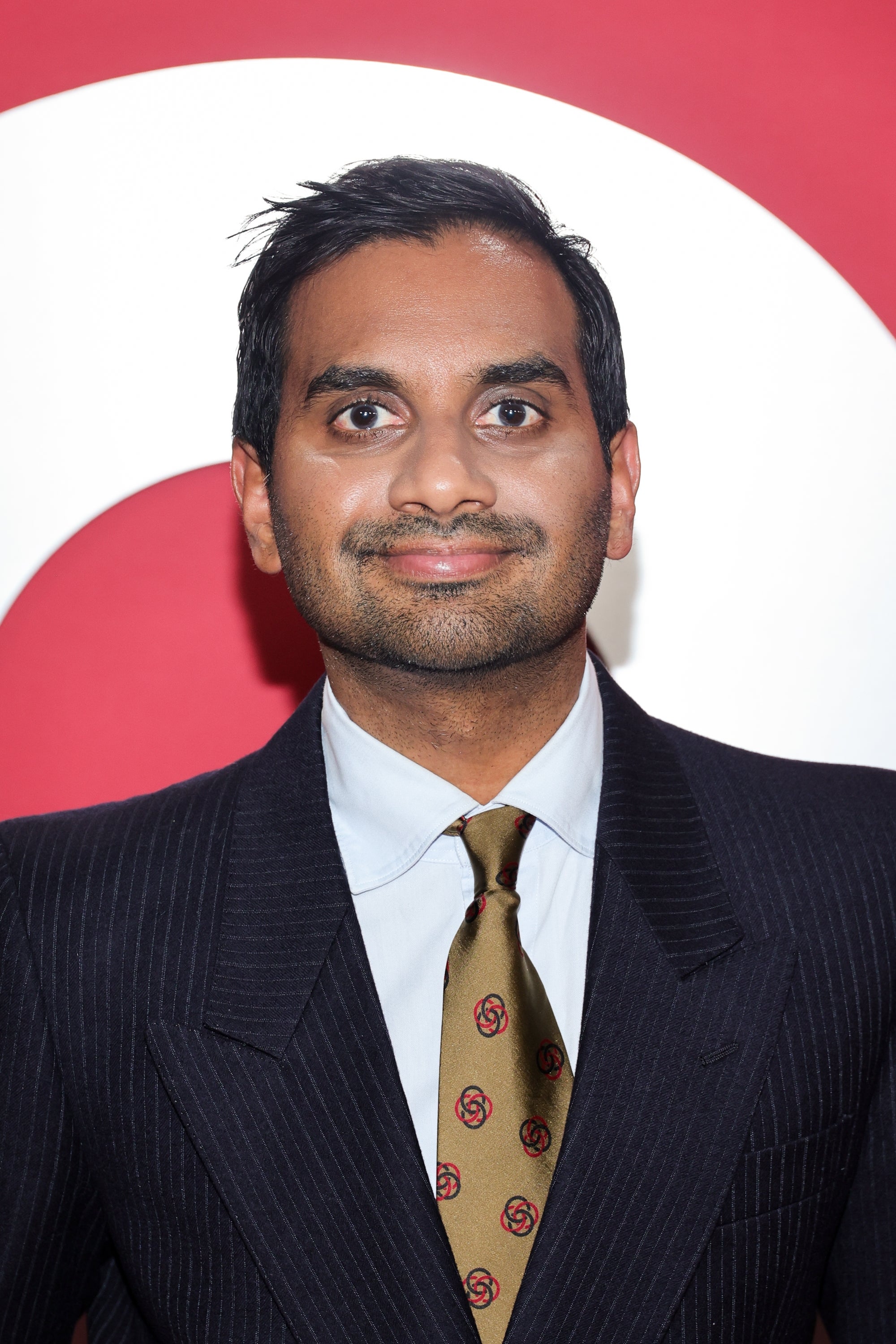 Aziz Ansari in a suit with a patterned tie on a red carpet event, standing against a bold backdrop