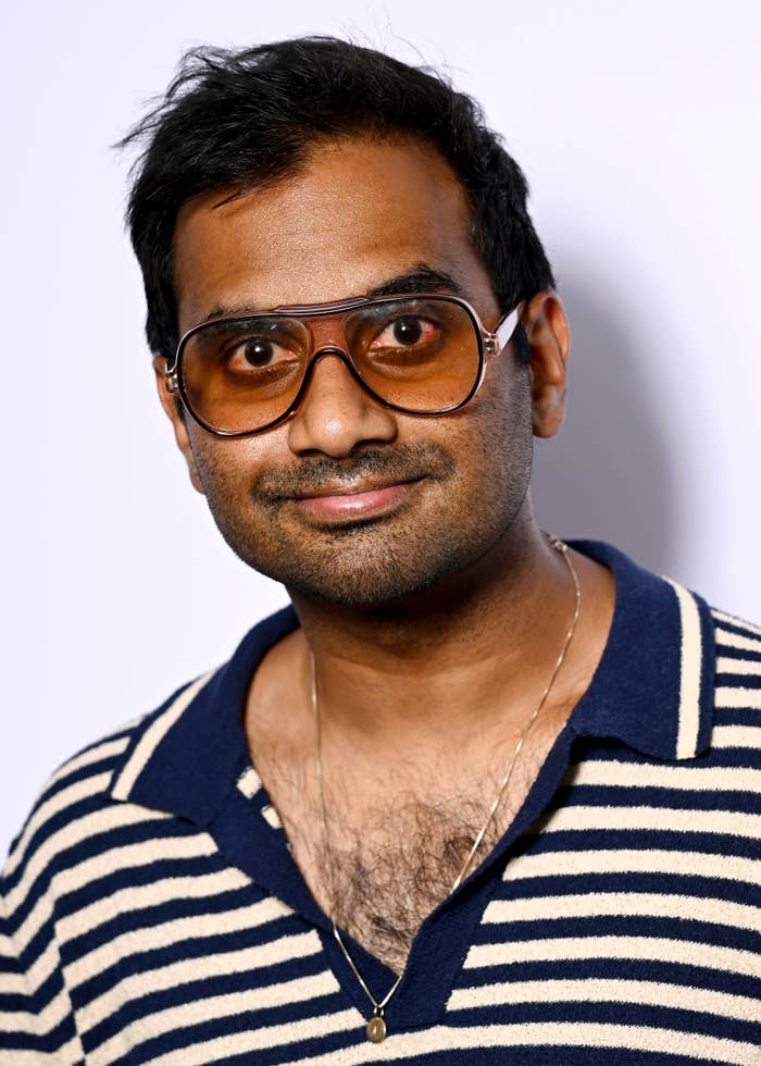 Aziz Ansari wearing large aviator glasses and a striped polo shirt, posing against a plain background