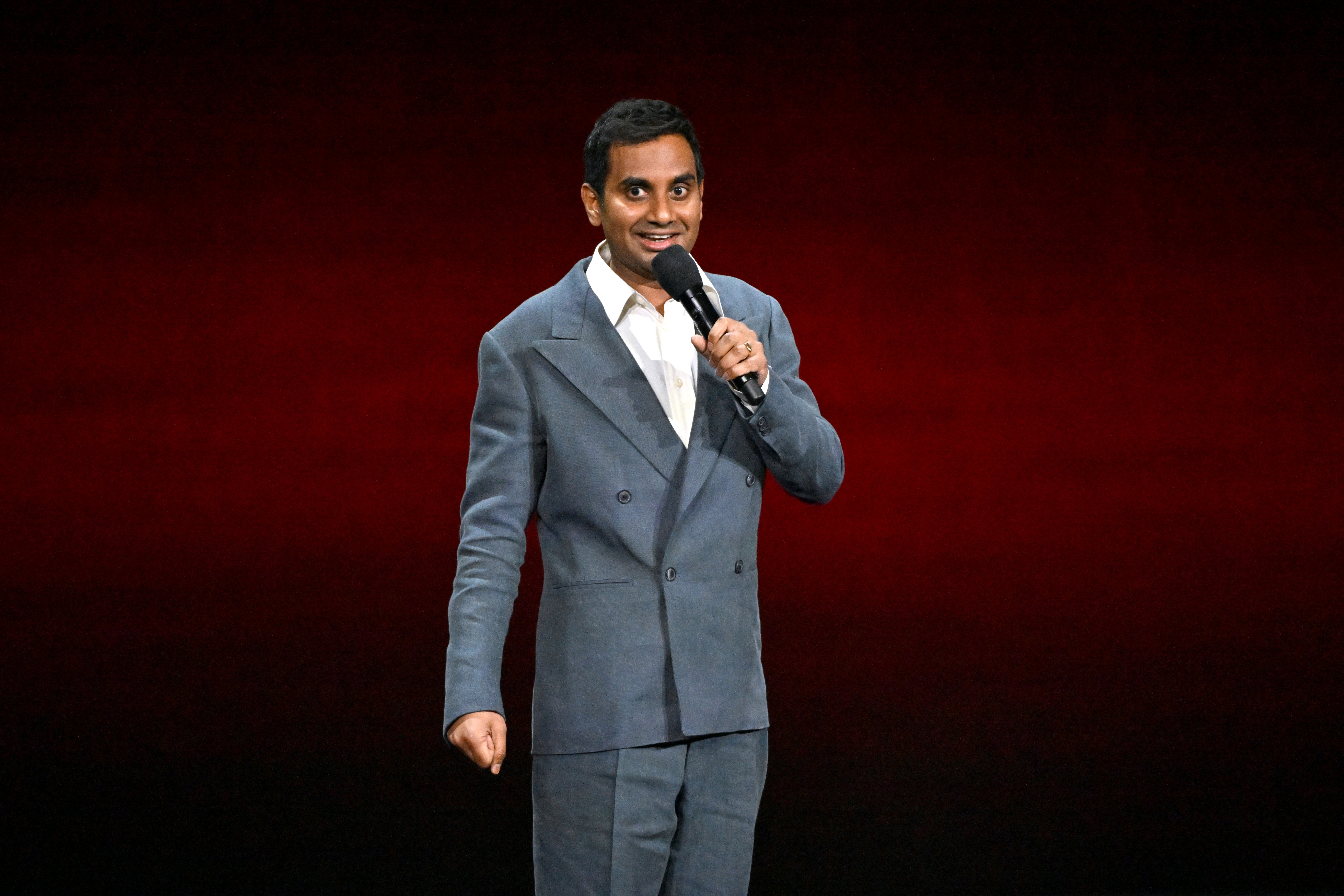 Aziz Ansari in a stylish double-breasted suit performs on stage, holding a microphone with a smile