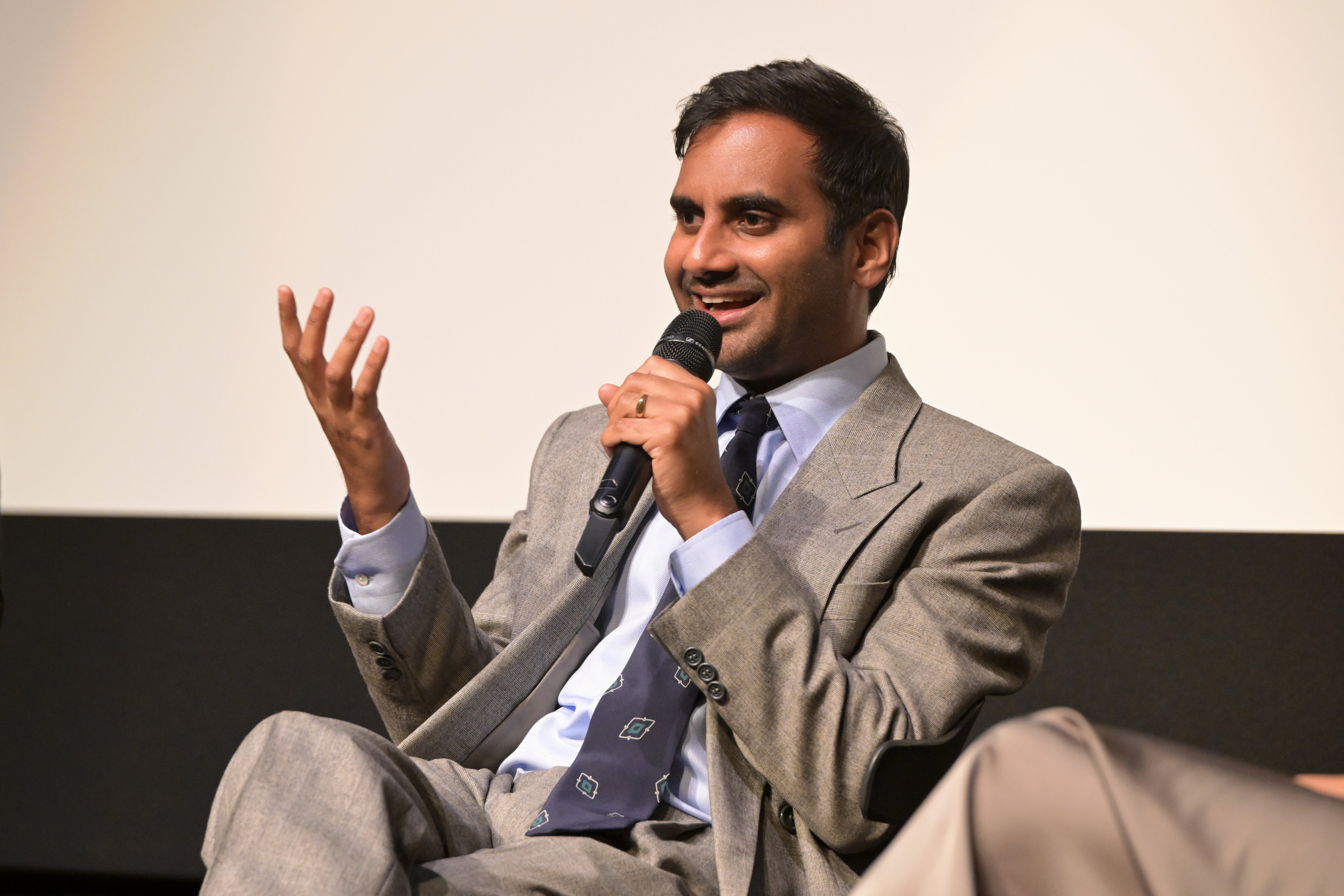 Aziz Ansari in a suit and tie, smiling and speaking into a microphone during an event