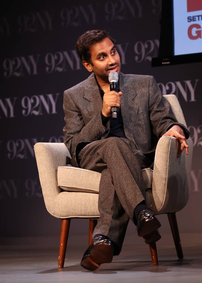Aziz Ansari  in a tailored suit sits on a stage armchair, holding a microphone, appearing engaged in a discussion or presentation