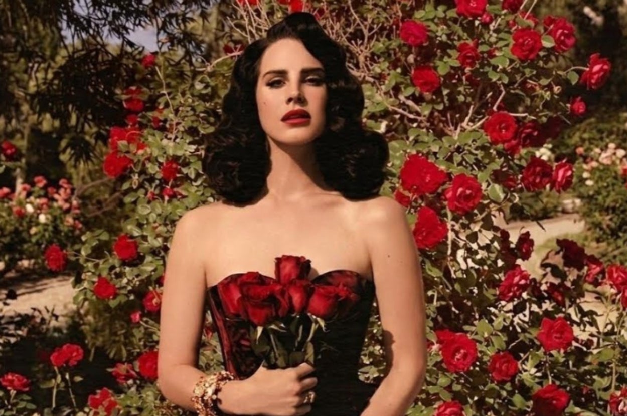 A woman with styled dark hair in a strapless dress holds red roses, standing amidst a background of blooming roses