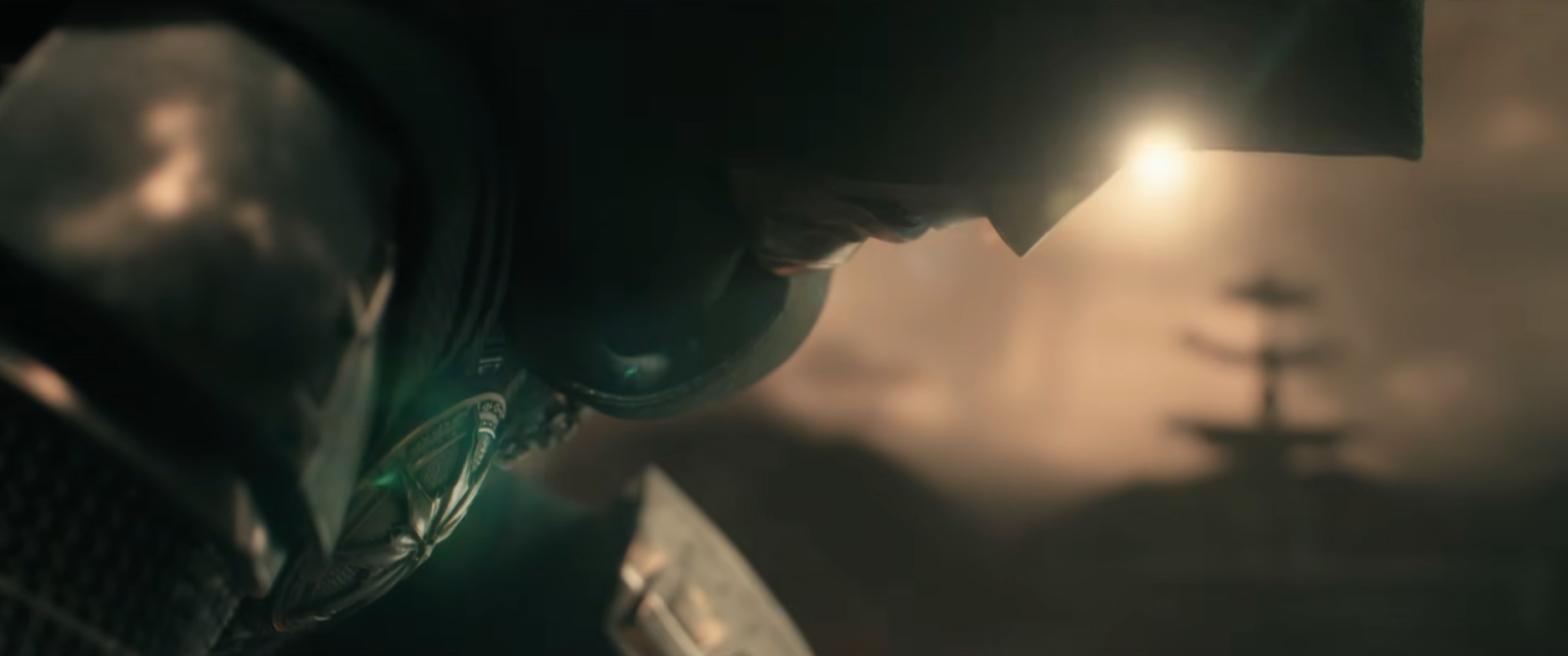 A warrior in metallic armor and hood bends forward in a dramatic scene, with light illuminating their face