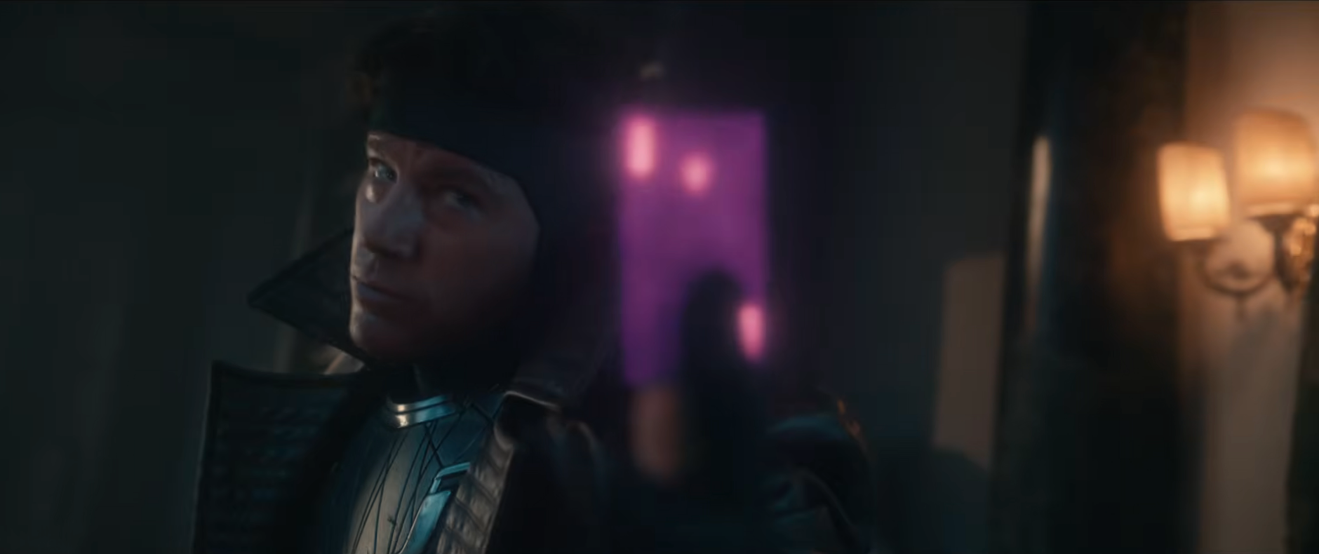 A person wearing a trench coat and headband looks sideways in a dimly lit room, with a blurred figure in the background
