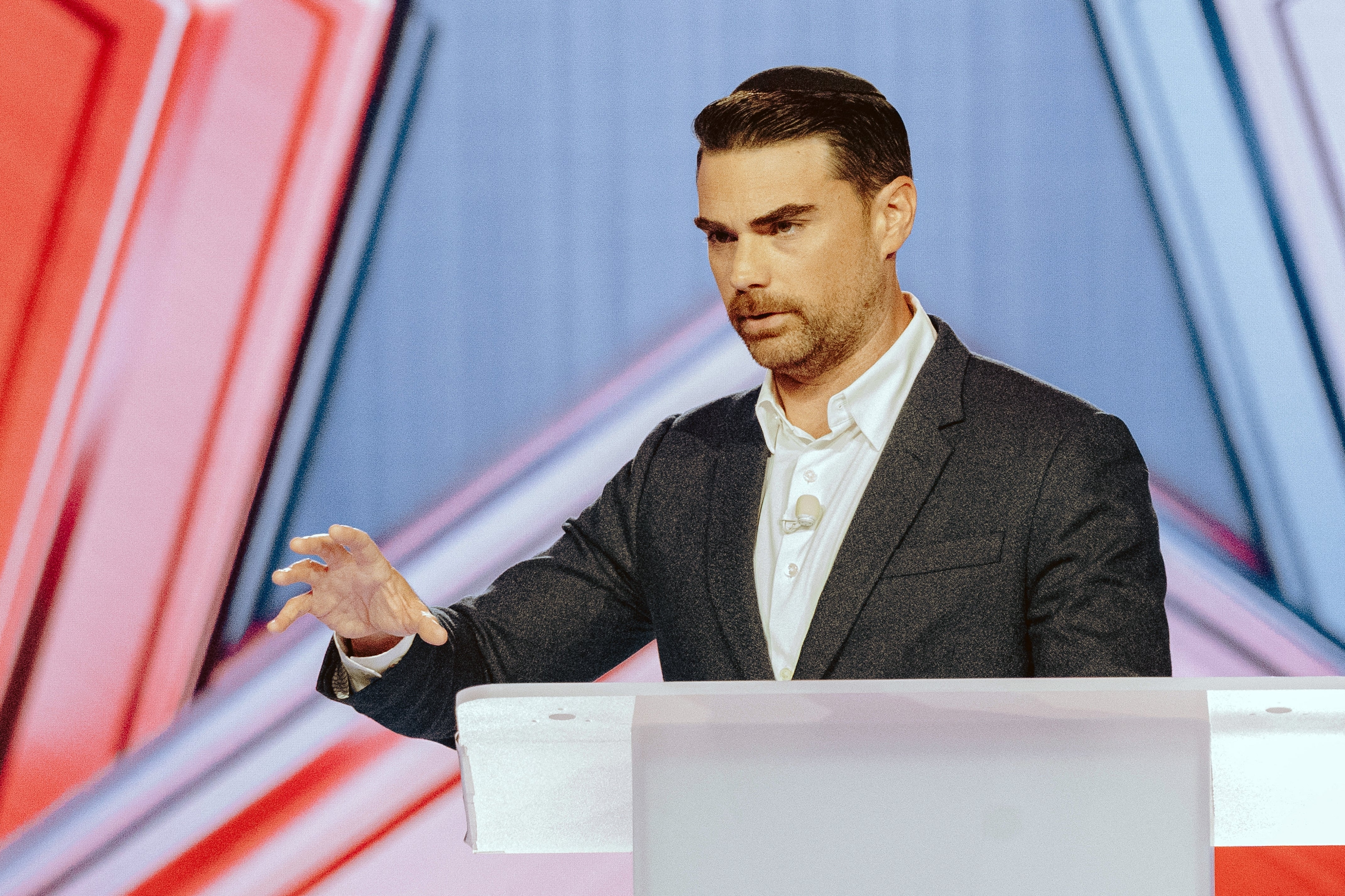 Man in a suit gestures while speaking at a podium with a geometric backdrop