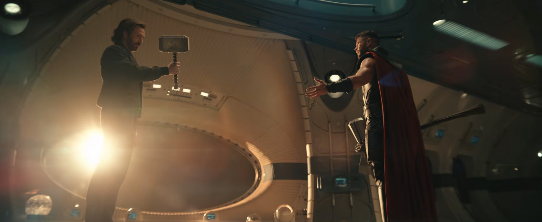 Two men stand facing each other in a futuristic room; one holds a hammer, while the other gestures with open arms, wearing a cape