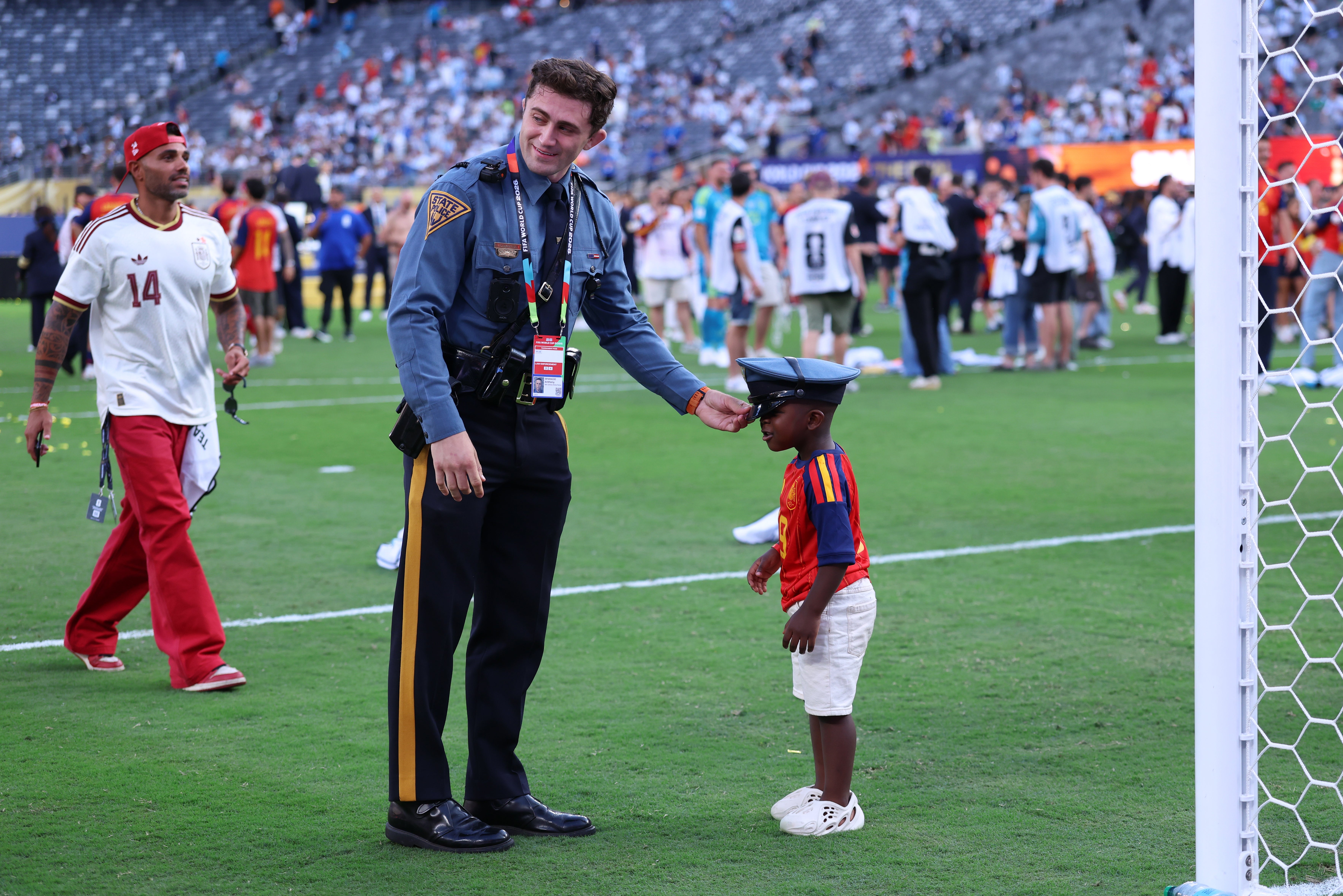 Police serviceman smiling and handing his chapeau to a young kid connected a sports field, with radical successful sports attire successful the background