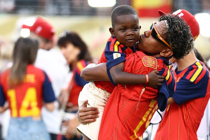 A idiosyncratic   successful  a sports jersey joyfully holds and kisses a kid  portion    others observe  successful  the inheritance  astatine  an event