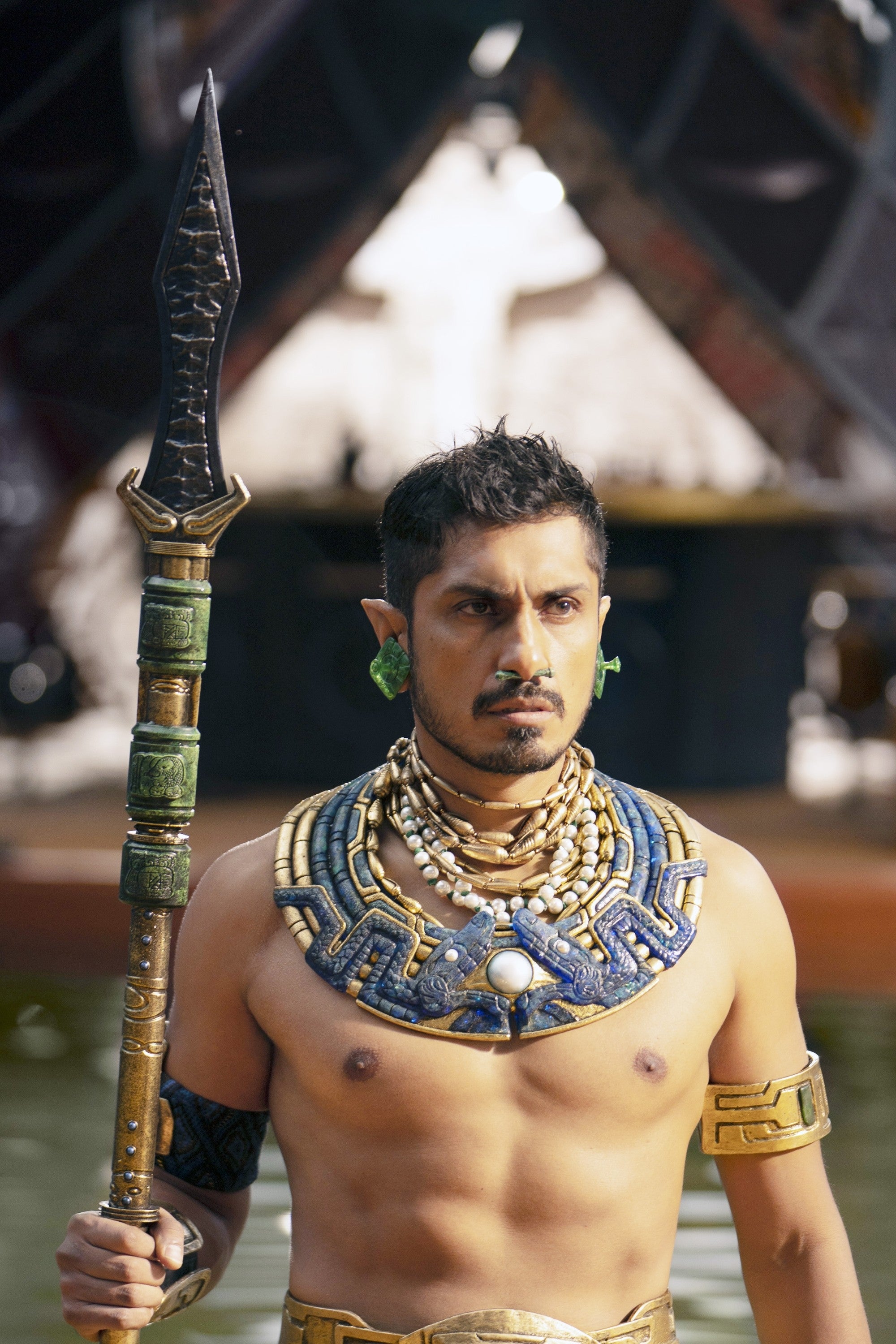 Person in a fantasy warrior costume with a spear and elaborate beaded jewelry, standing in a dramatic pose