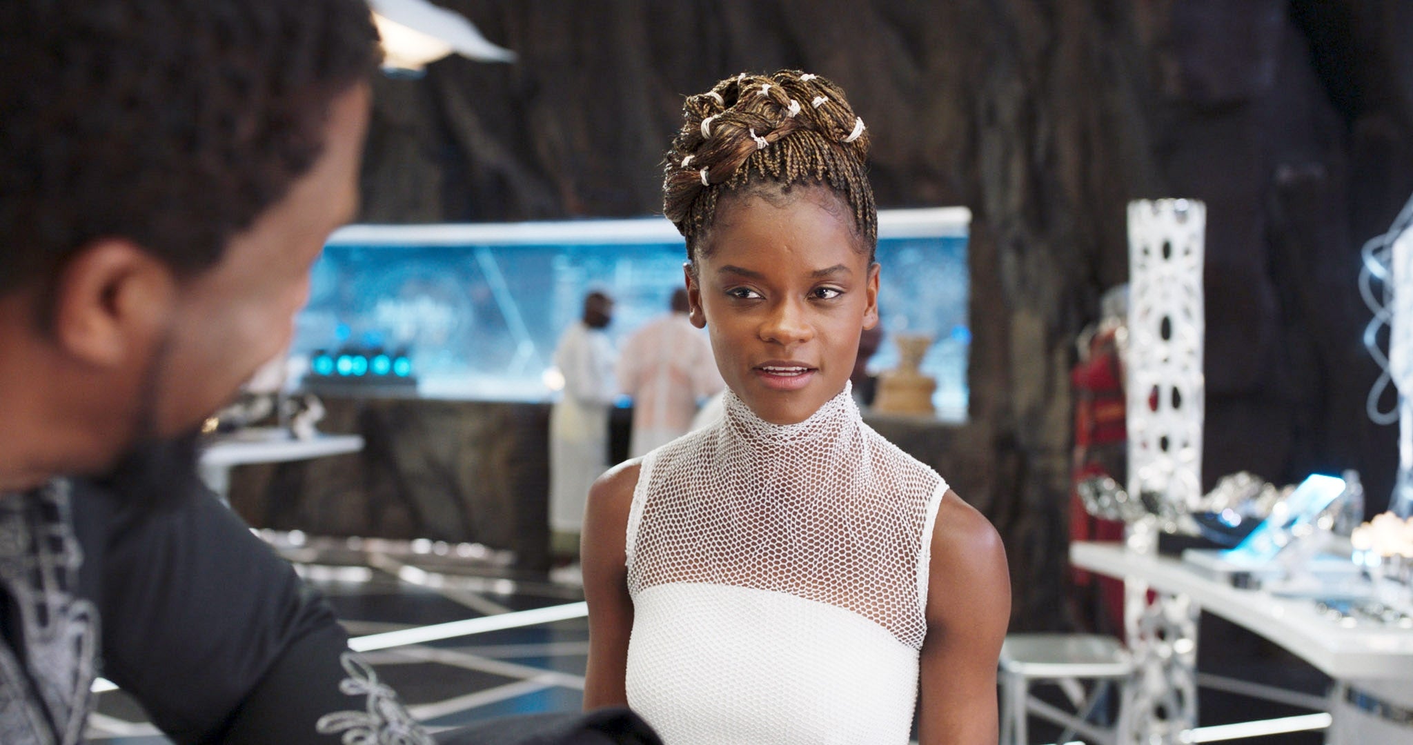 A person in a futuristic lab setting talks to someone. They wear a sleeveless, high-neck dress with a mesh upper part