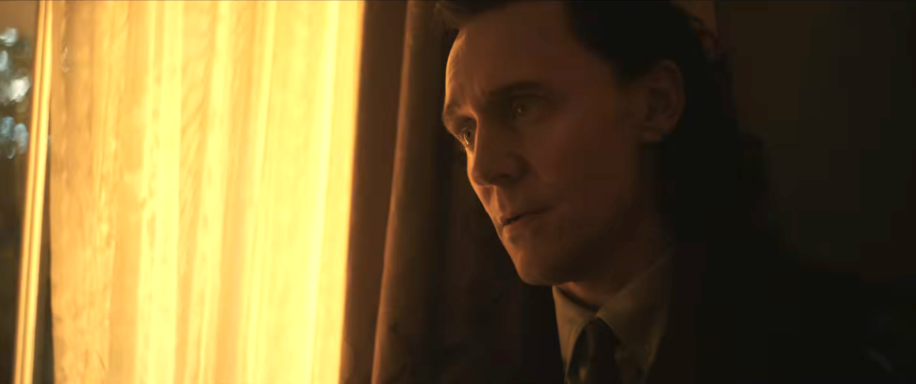 A person with long hair in a dimly lit setting looks contemplatively out of a window. They are wearing a suit