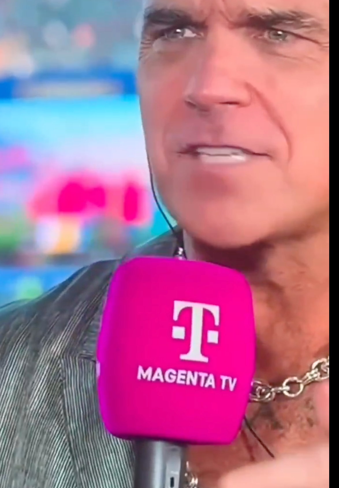 Orang yang berbicara ke mikrofon magenta berlogo, mengenakan jaket bergaris-garis dan kalung rantai