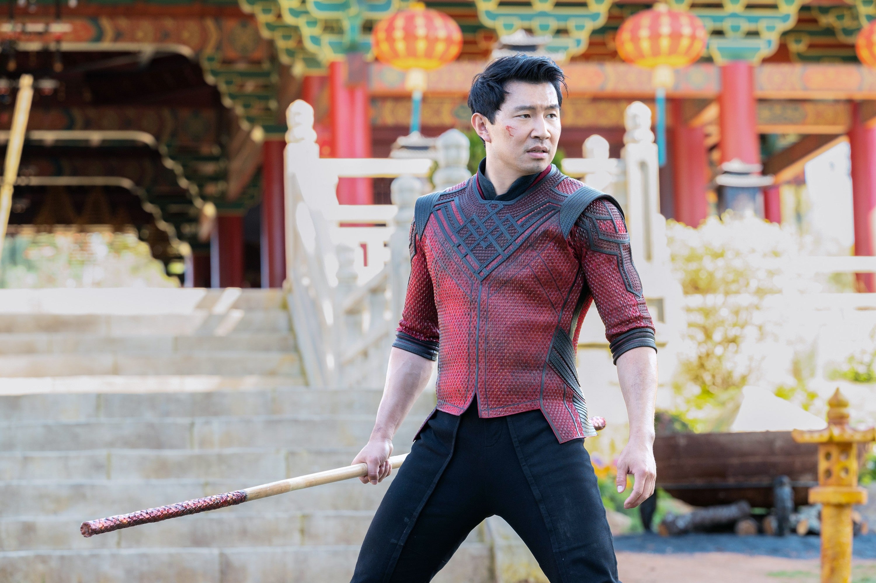 Person in a red and black, detailed warrior costume holds a staff, standing in front of ornate architecture with lanterns