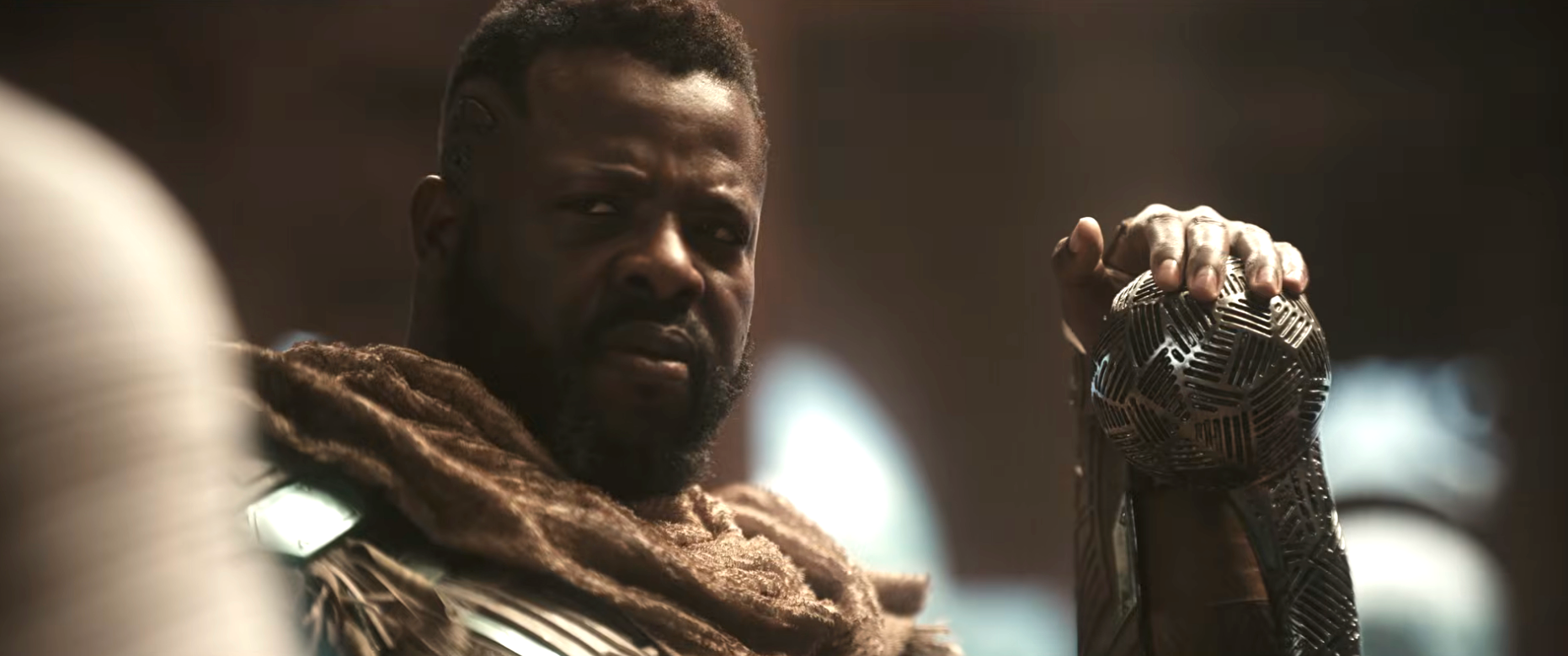 Person wearing textured armor and a detailed cloak, posing dramatically while holding a metal sphere