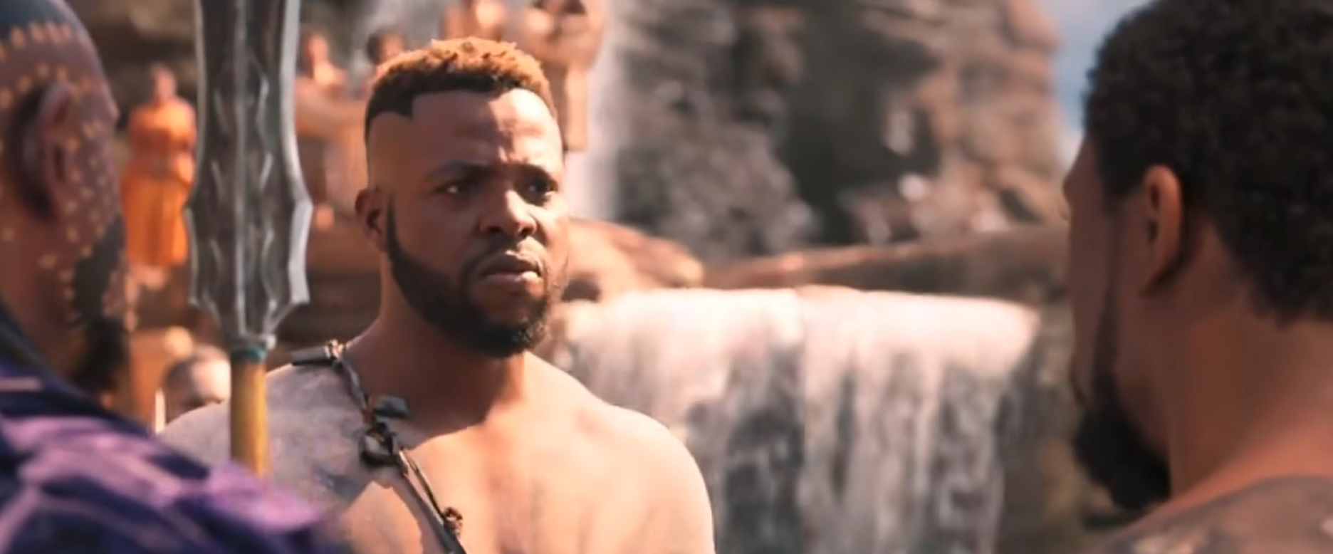 Scene from a movie with three characters, one speaking to another near a waterfall. They are dressed in tribal-style outfits