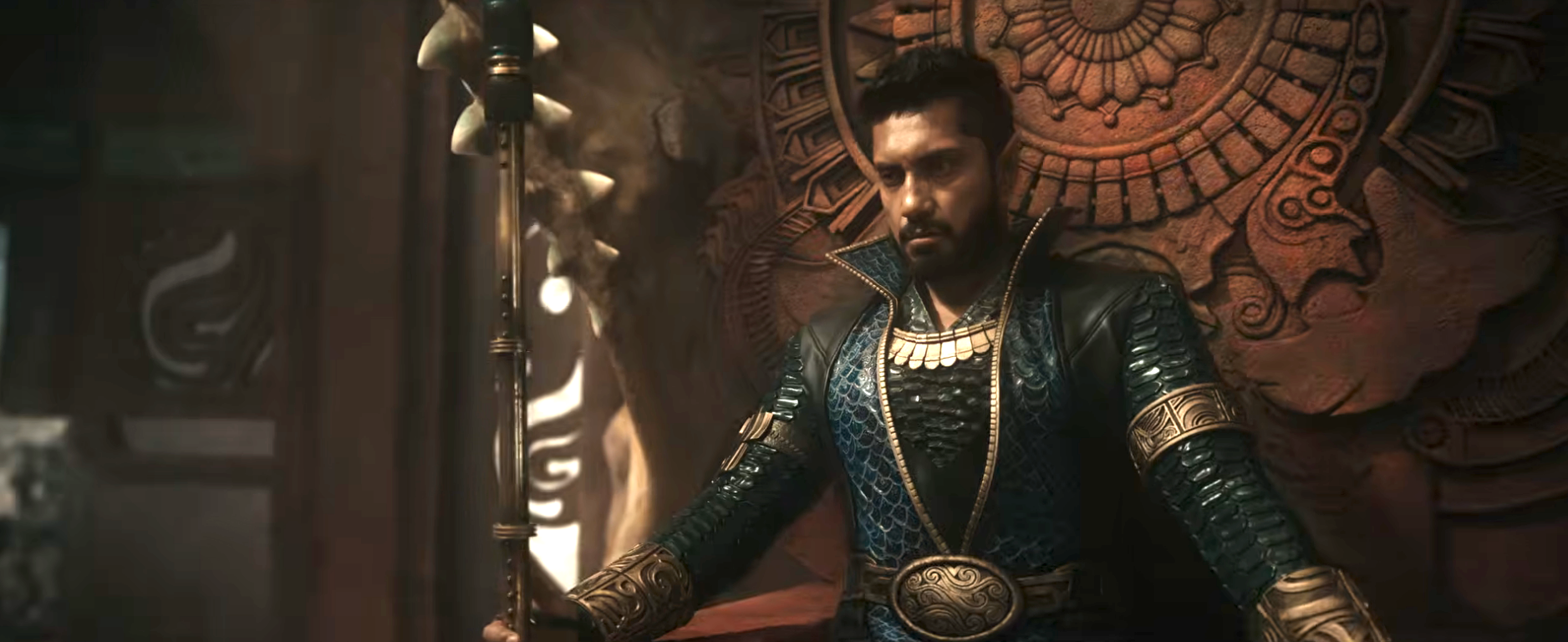 Person dressed in intricate, ornate attire, seated on a throne, holding a staff in a dramatic, regal setting