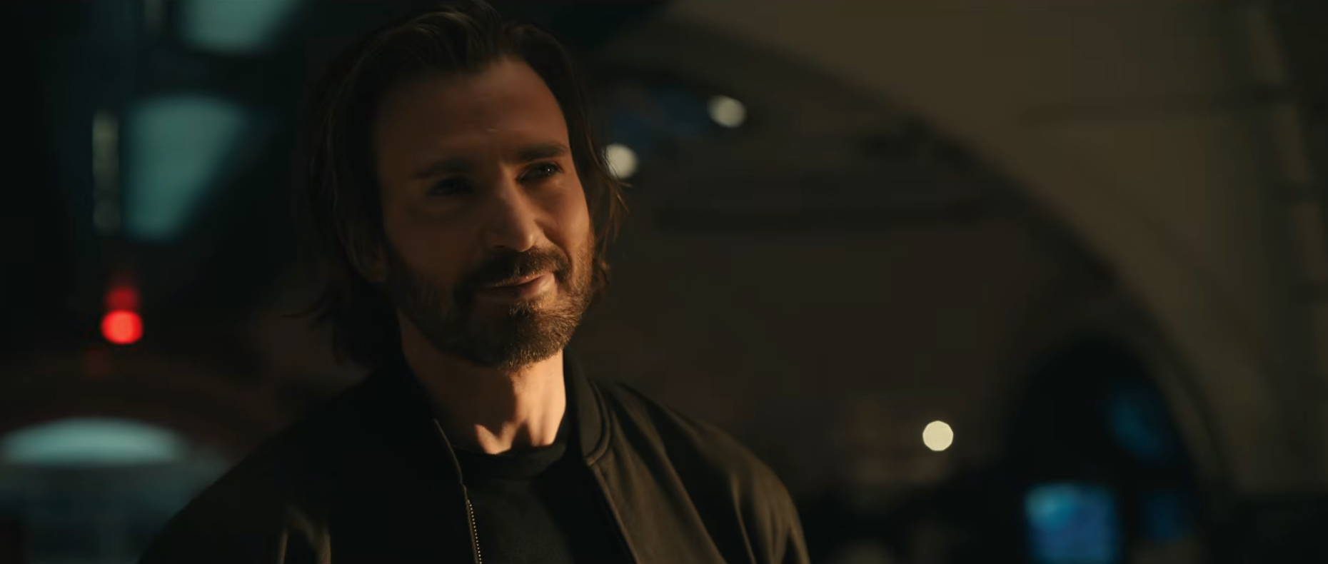 Man with medium-length hair and a beard stands indoors wearing a dark jacket, appearing thoughtful
