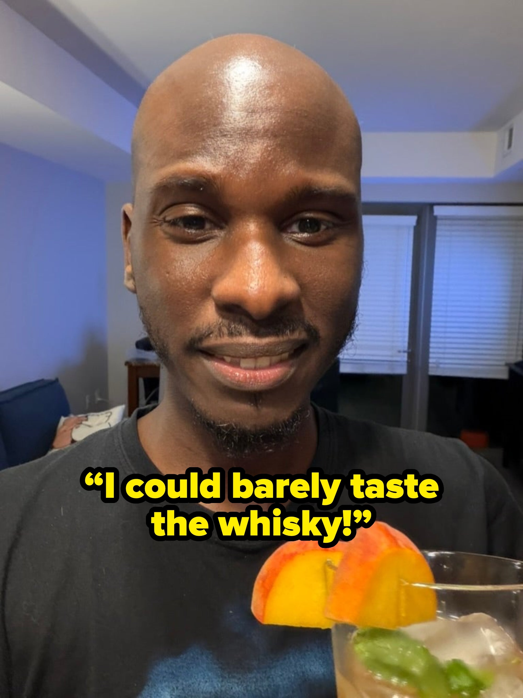 A person smiling while holding a glass with ice, peach slices, and green garnish, suggesting a refreshing beverage or cocktail