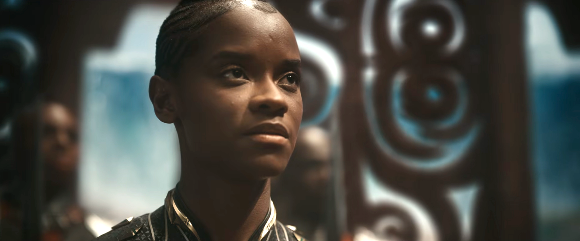 Person with braided hair in ornate room, wearing a patterned top with a high collar, looking forward with a focused expression