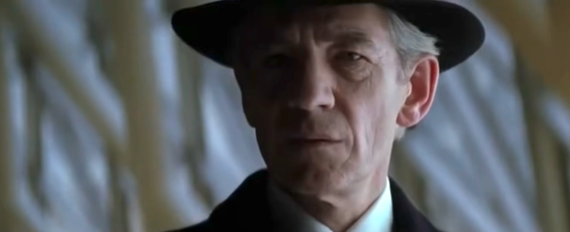 An older person in a classic suit and fedora hat, with a neutral expression, stands before an abstract background