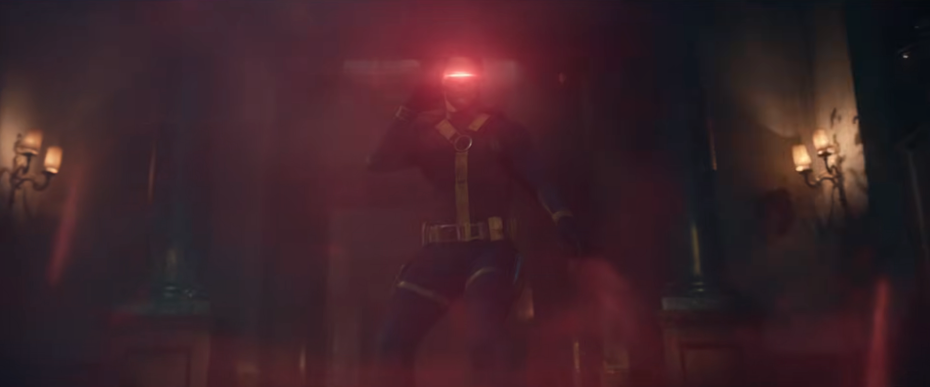 A superhero figure emits a bright red light from their visor in a dimly lit room, standing in a combat-ready pose