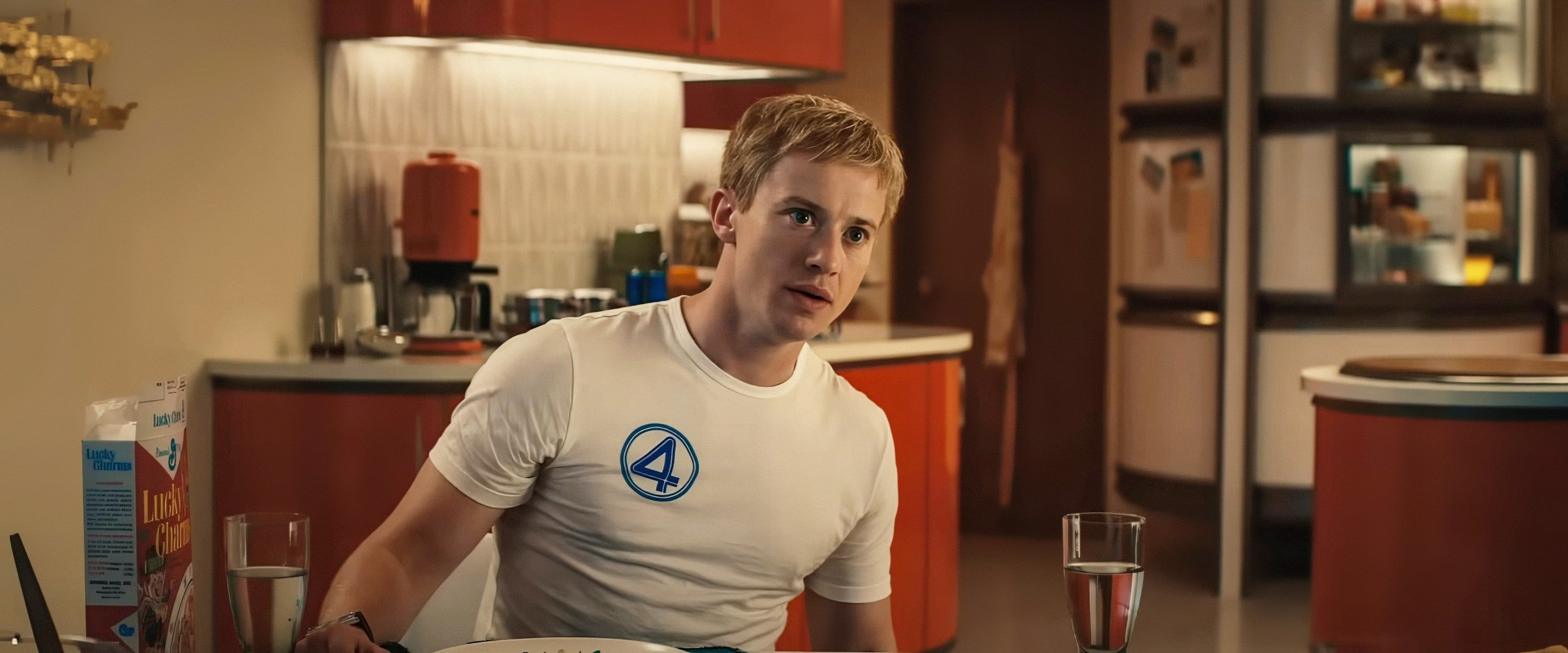 Person seated at a kitchen table, wearing a white shirt with a blue "4" logo, looking attentive. Kitchen items are visible in the background