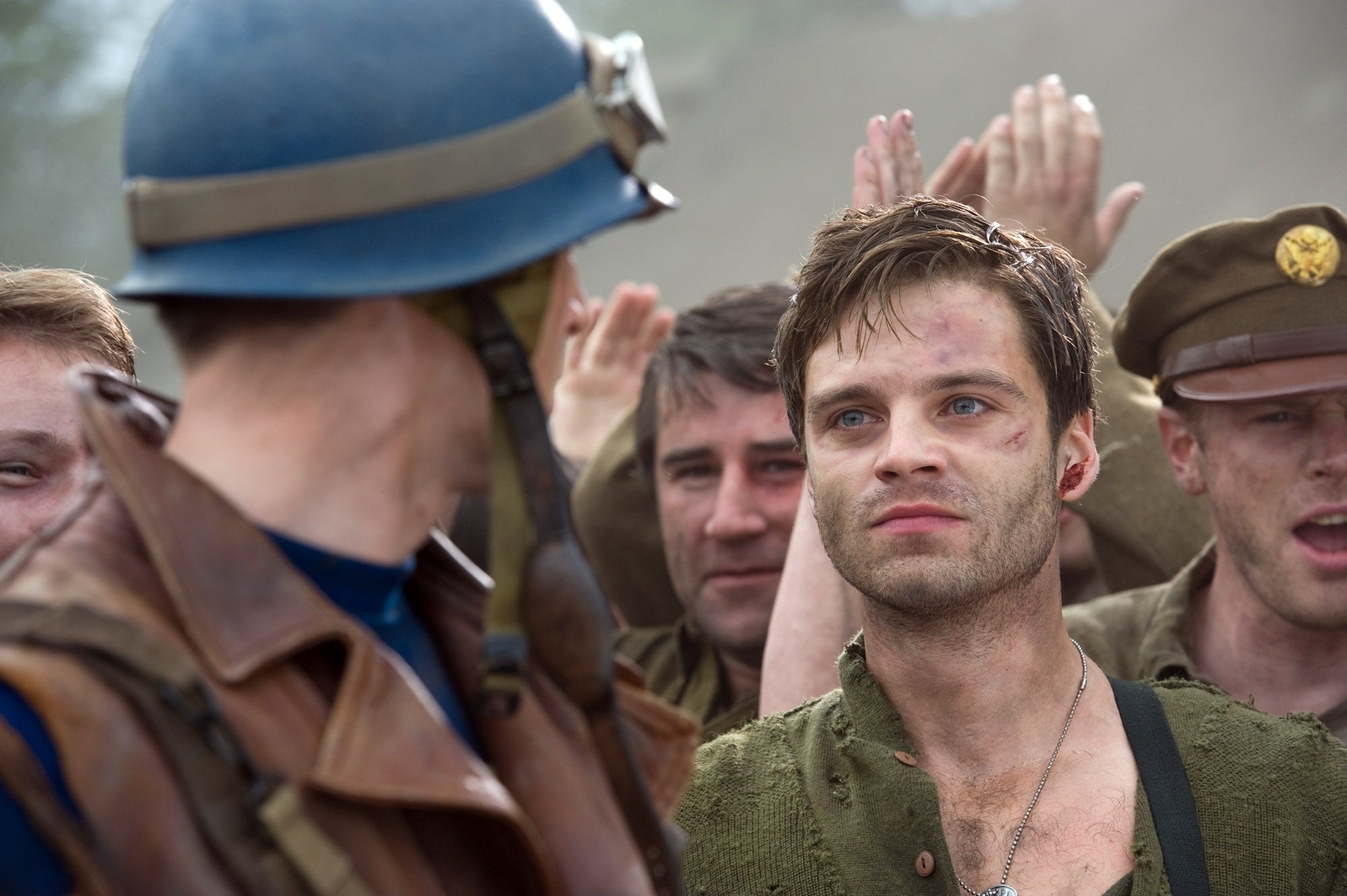 Two men in military attire during WWII-era scene, one in a helmet facing away, the other looking distressed with comrades behind