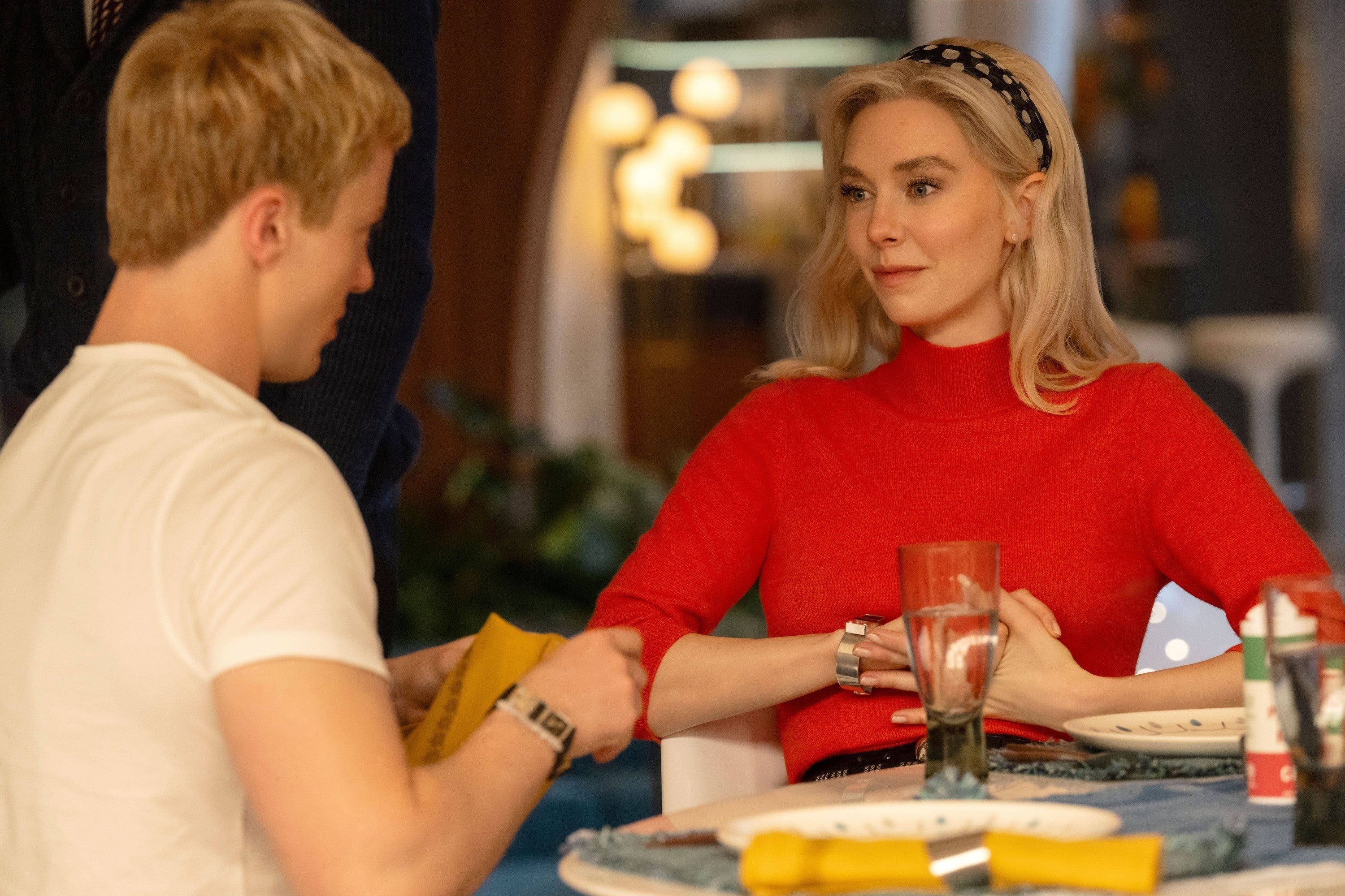 Two people sit at a dining table, engaged in conversation. One wears a red top with a headband, the other in a white shirt