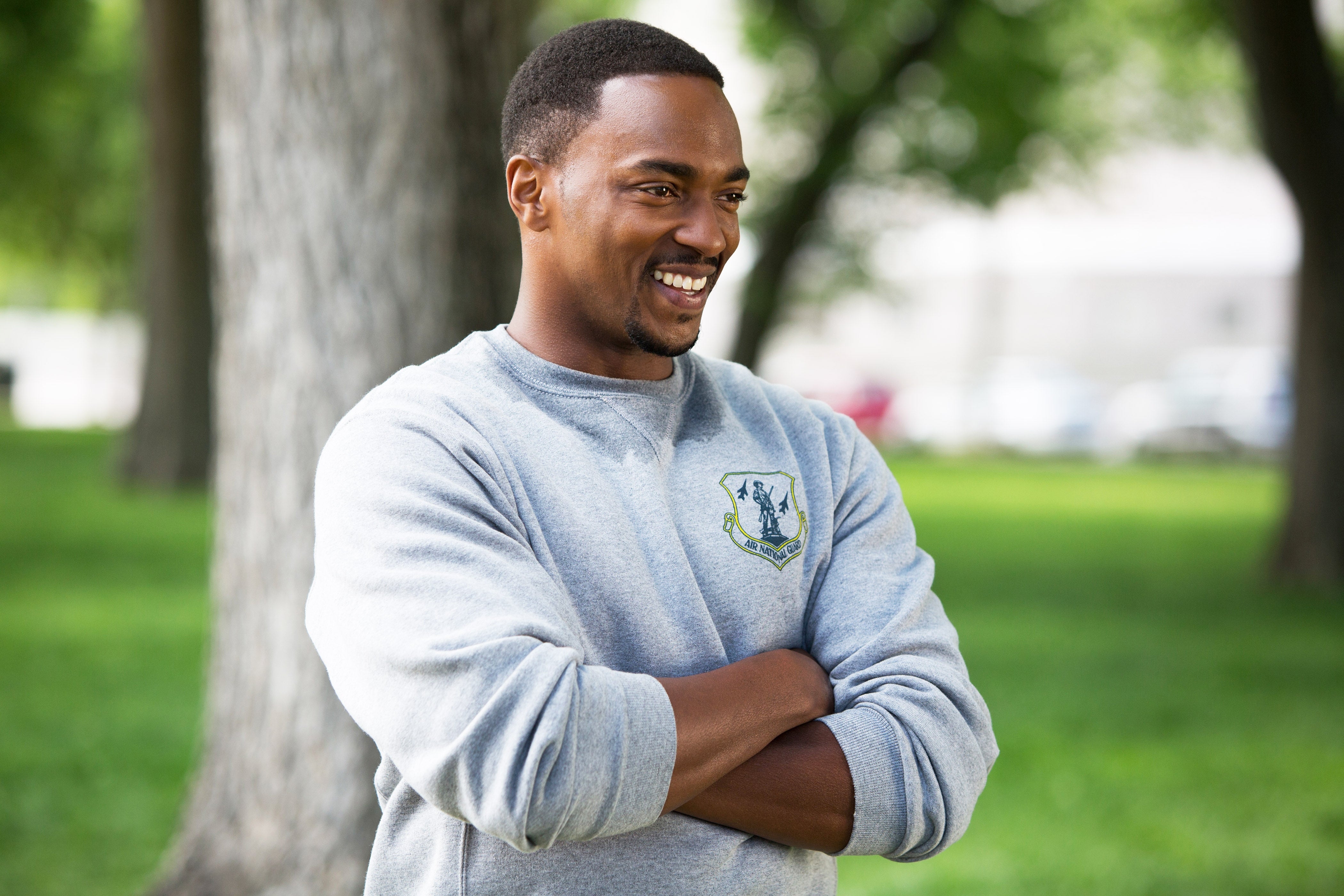 Person outdoors, smiling, arms crossed, wearing a casual sweatshirt with an emblem, standing in a park with trees in the background
