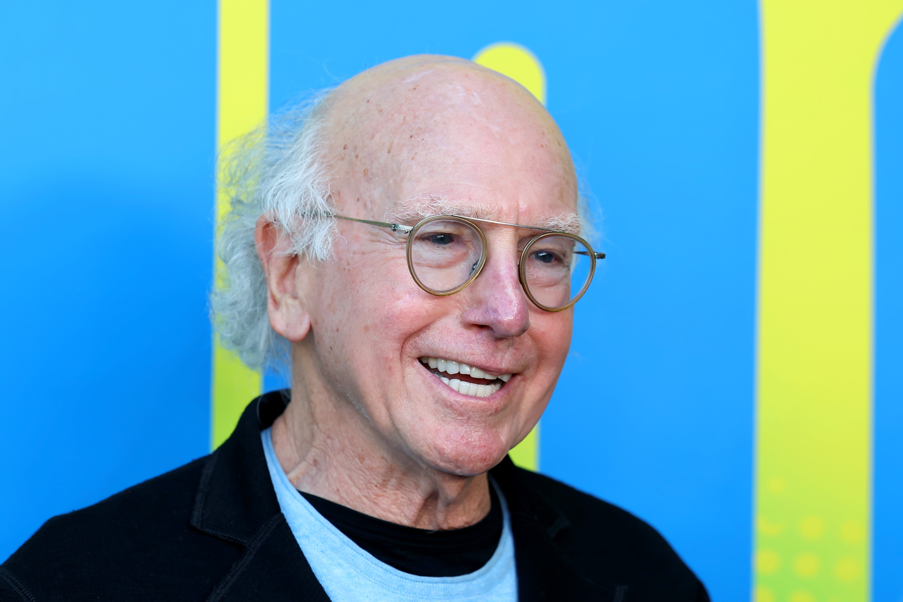 Smiling older man with glasses and a bald head, wearing a casual black jacket over a light shirt, with a bright blue background behind him
