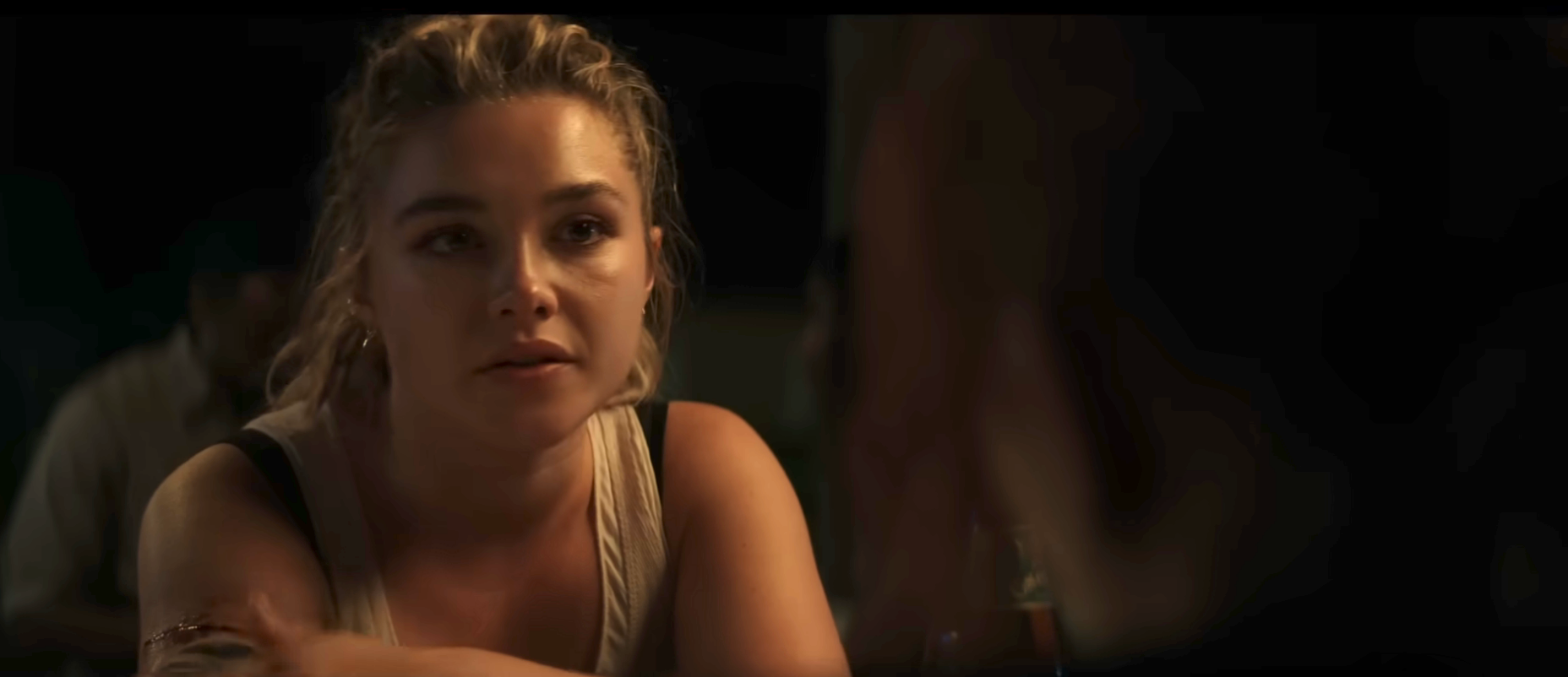 Person with pulled-back hair, wearing a sleeveless top, appears to be engaged in a conversation, seated at a table in a dimly lit setting