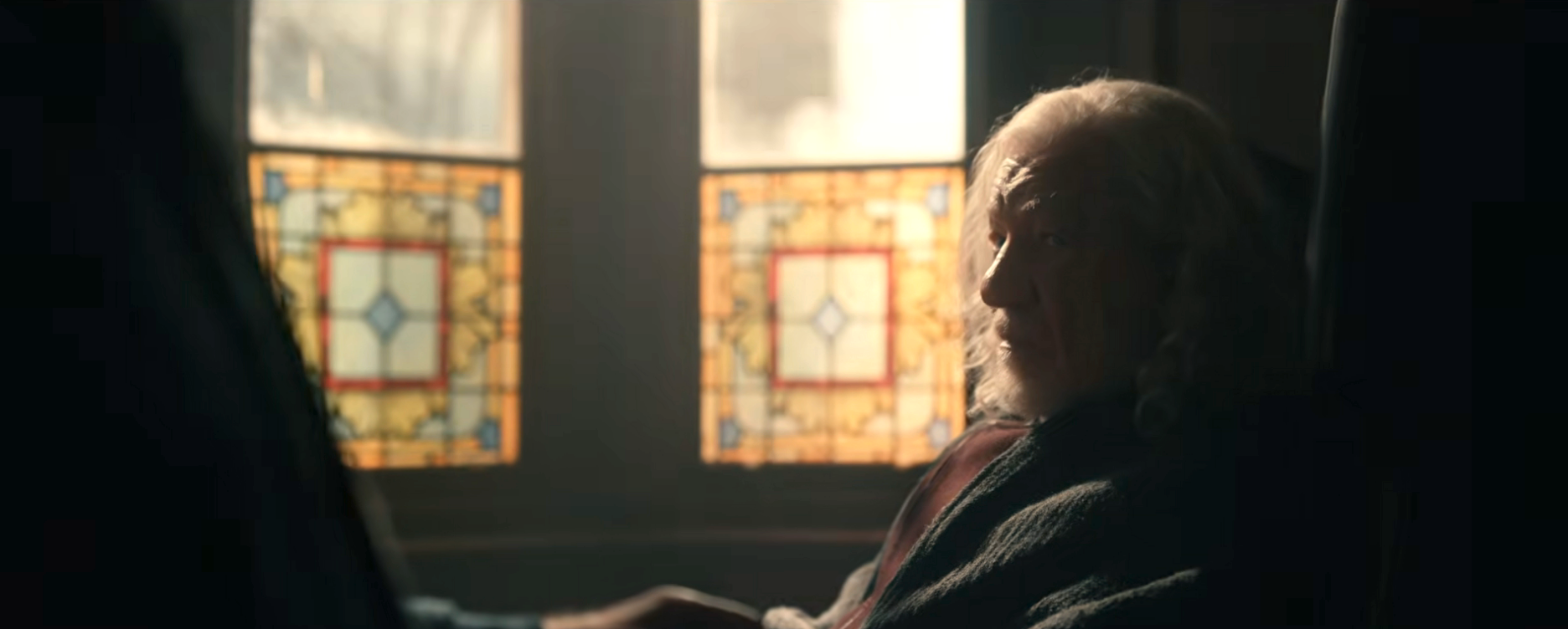 Elderly person sitting thoughtfully beside a stained glass window with a gentle light illuminating the scene