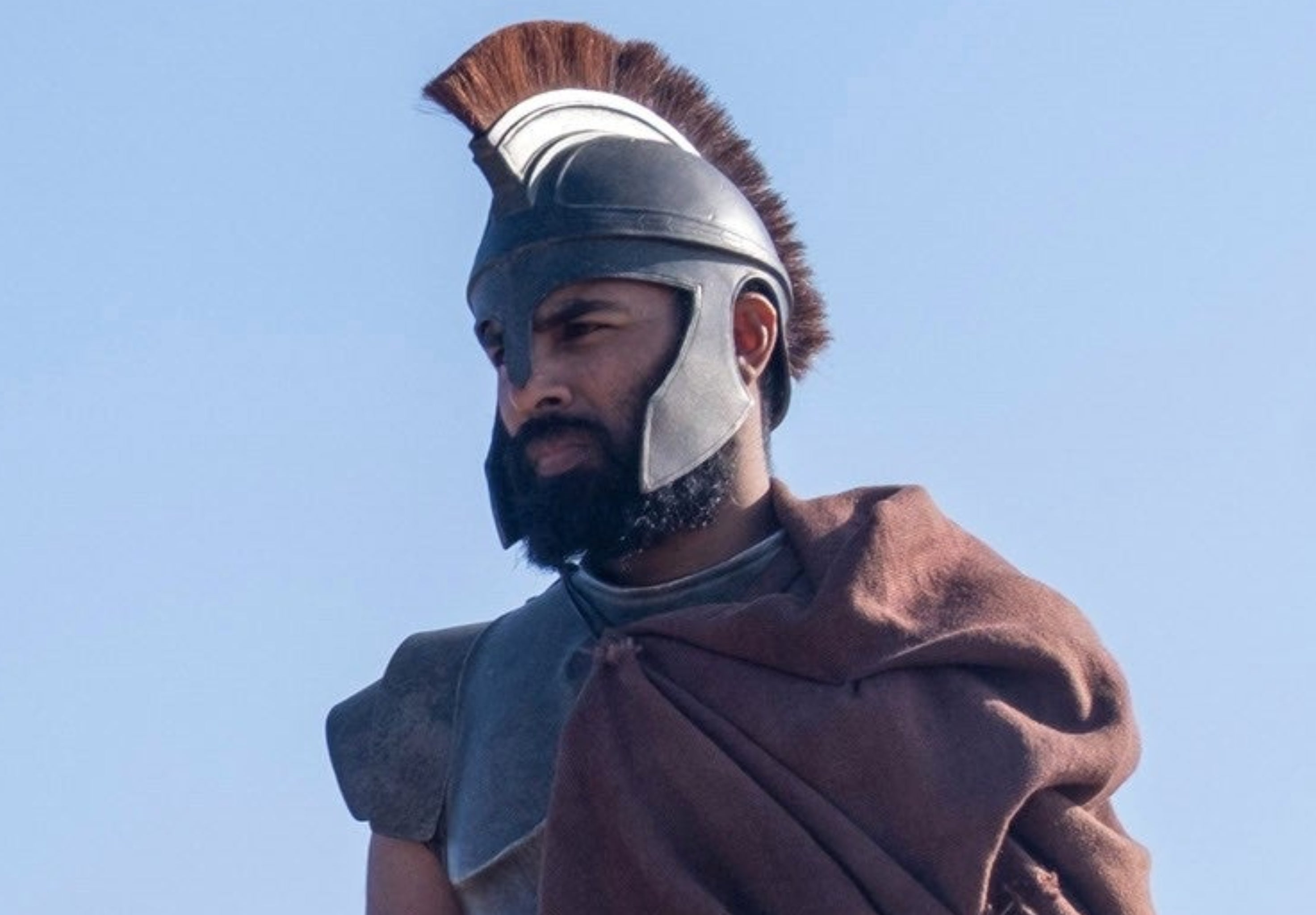 Three actors in ancient warrior costumes with helmets and armor, standing on a sandy landscape, possibly on a movie set