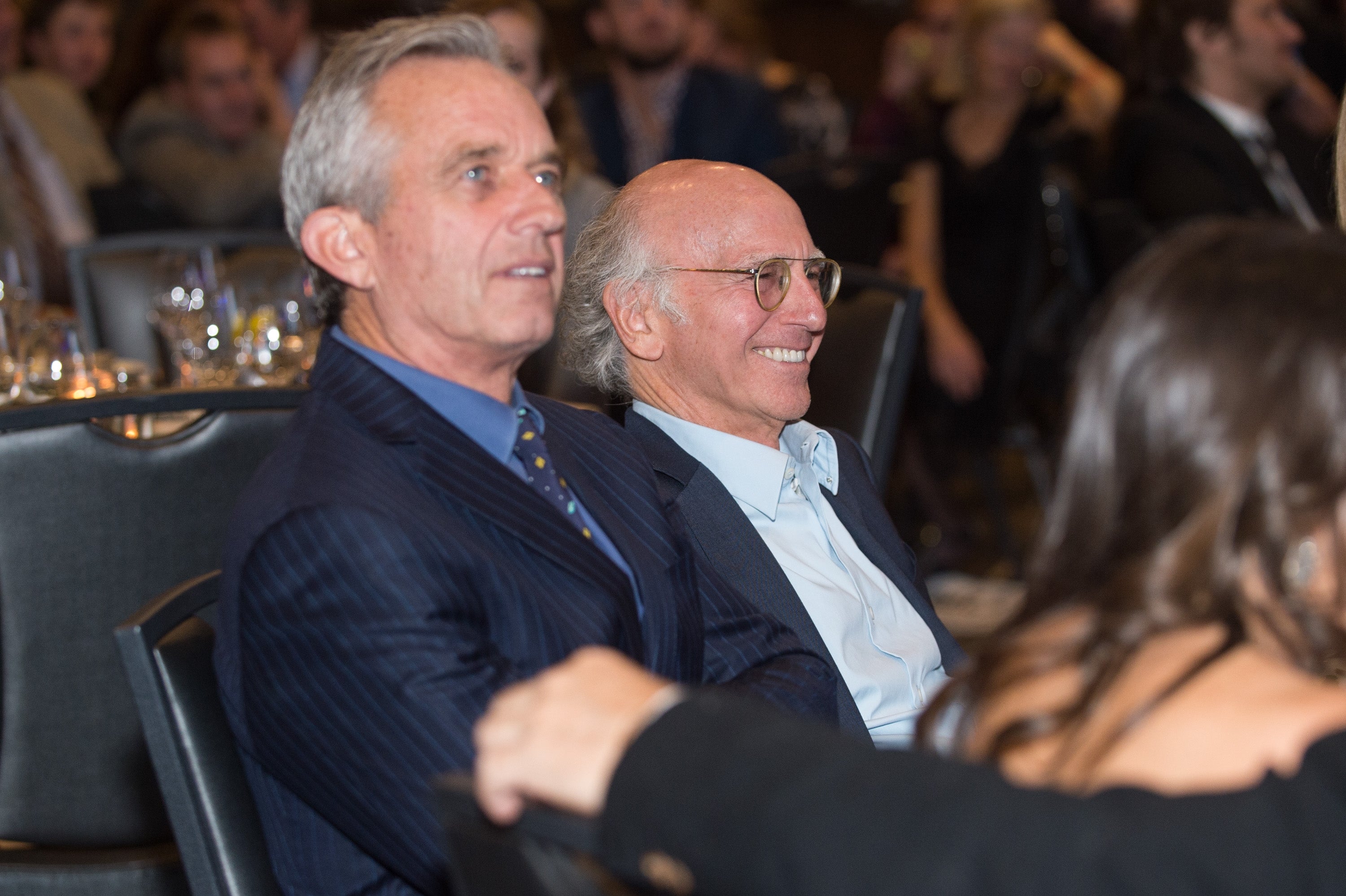 Two people seated at an event, smiling and wearing suits, surrounded by others