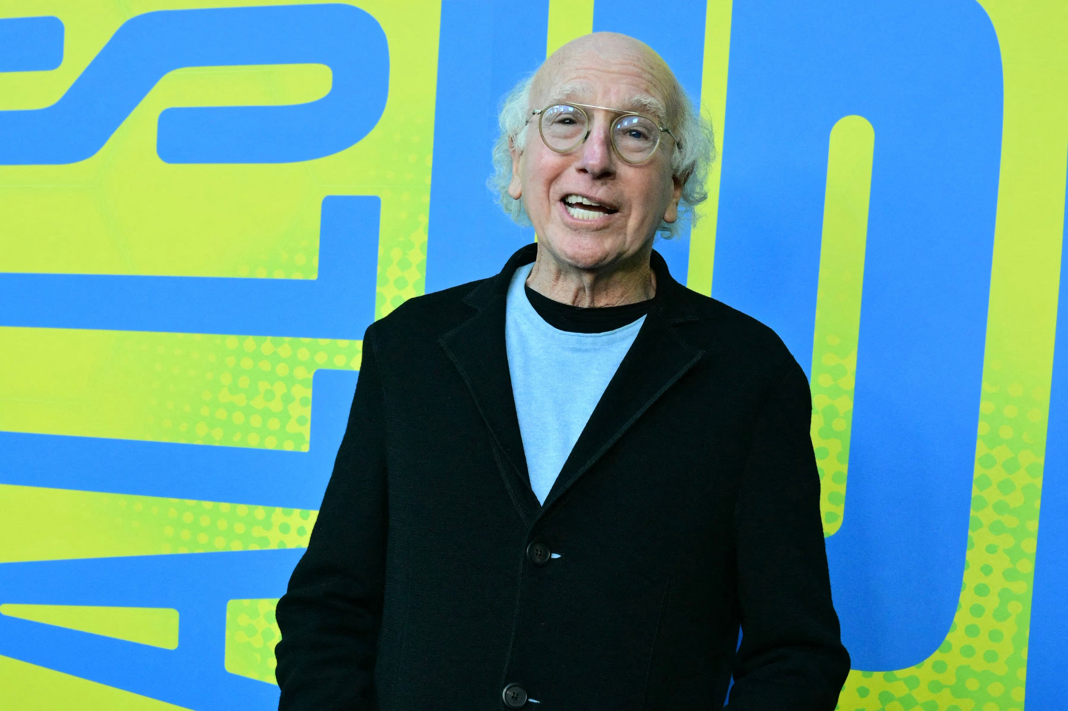 Person wearing glasses and a black jacket over a light shirt, smiling against a large graphic backdrop